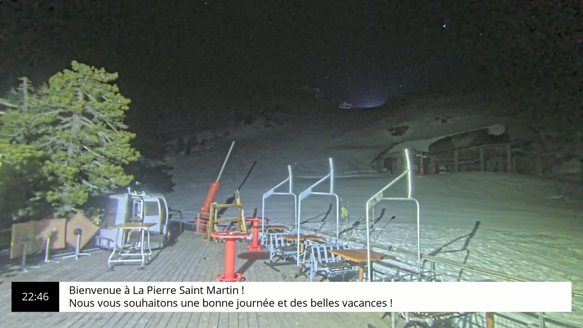 A nighttime view of a snow-covered ski slope with a chairlift, tables, and chairs on a wooden deck under a clear, starry sky.