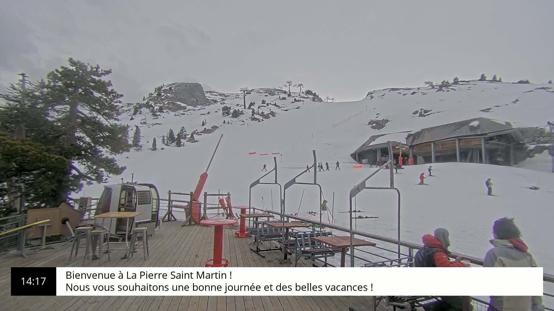 A snow-covered ski slope with several skiers and a chairlift, viewed from a wooden deck with outdoor seating under an overcast sky.