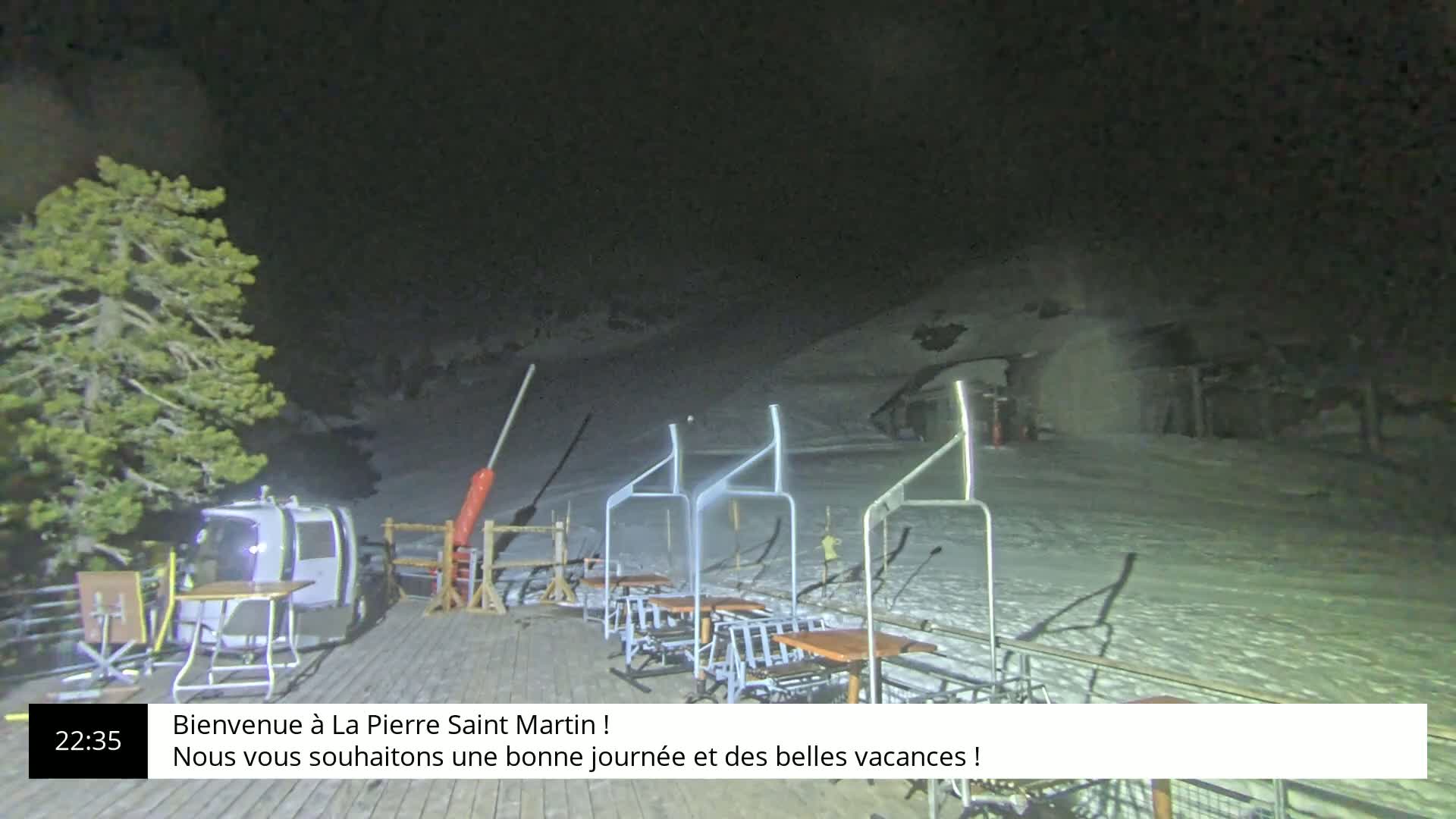 A nighttime view of a snow-covered ski slope with a chairlift and several tables and chairs on a wooden deck, under a dark sky.