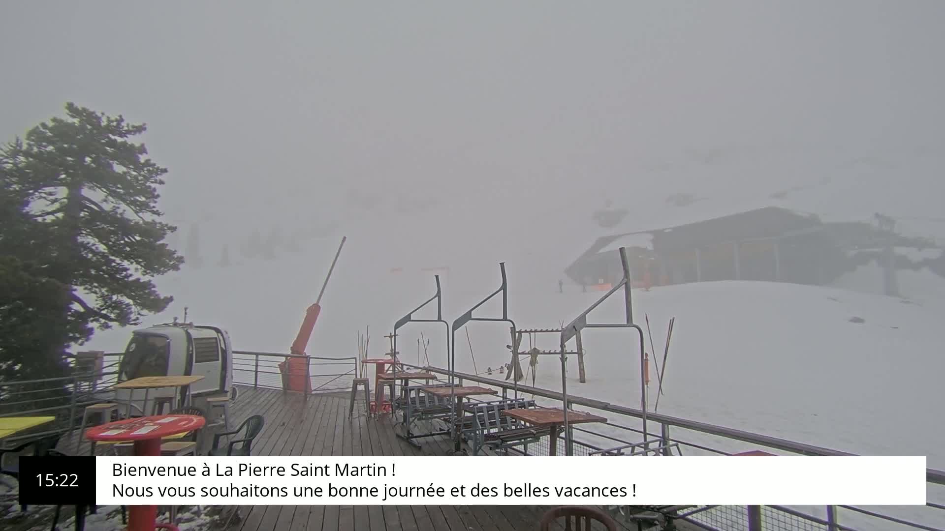 A snow-covered ski resort is shrouded in thick fog, with a chairlift and outdoor seating visible.