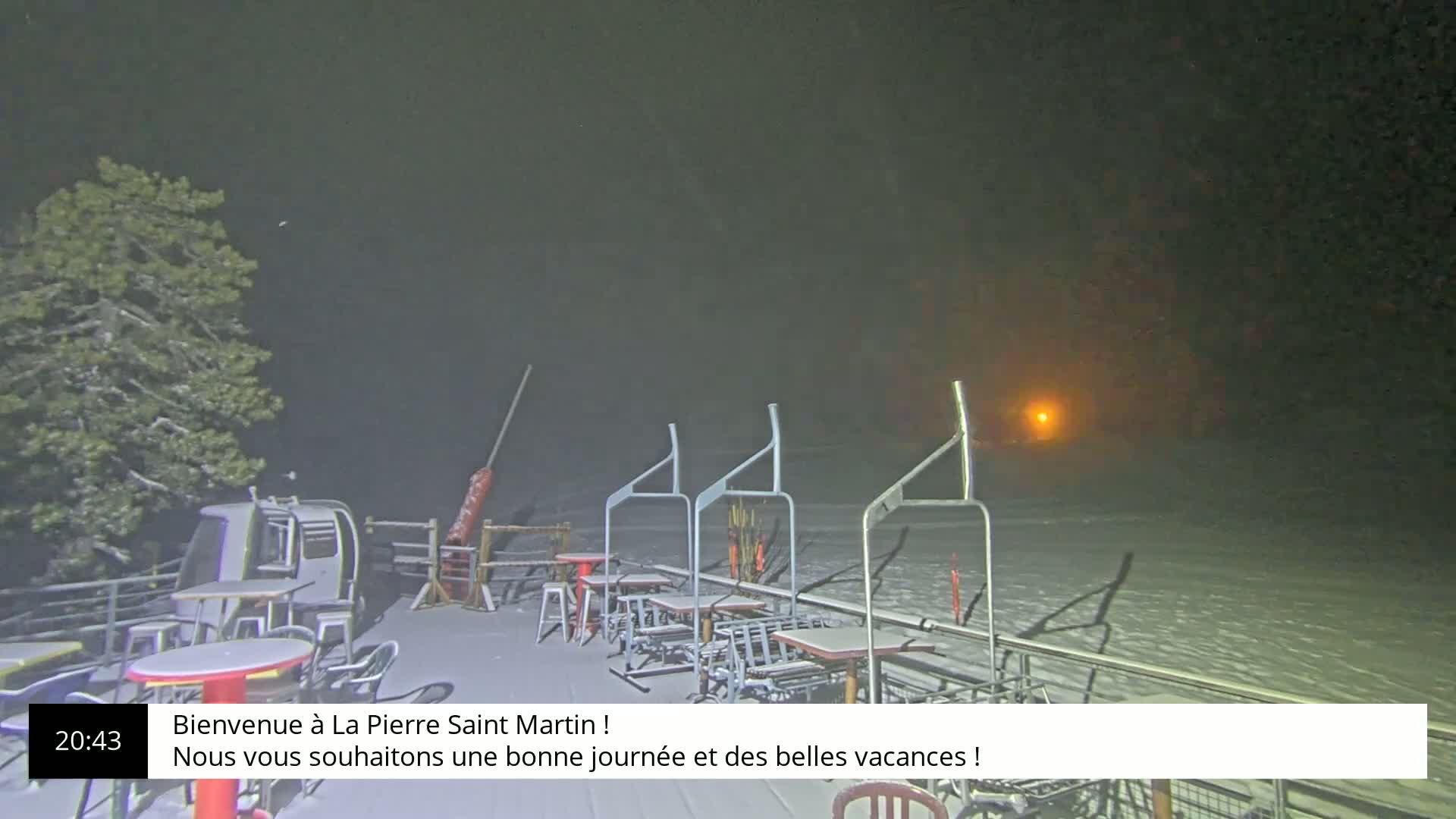 A snow-covered outdoor patio with empty tables and chairs sits under a hazy, dark night sky.