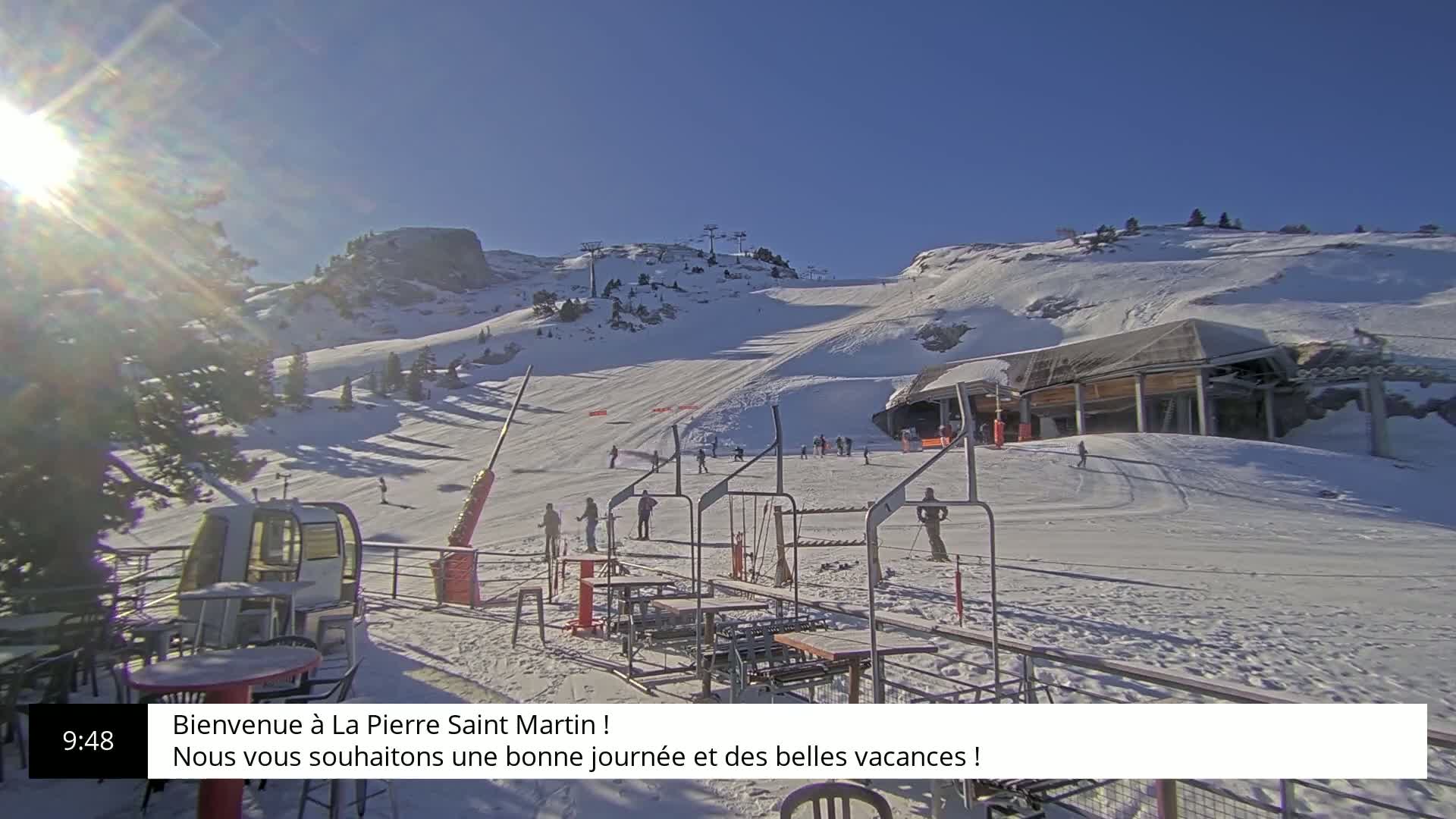 A sunny, snow-covered ski slope with skiers and a lift station is visible from a patio with outdoor seating.