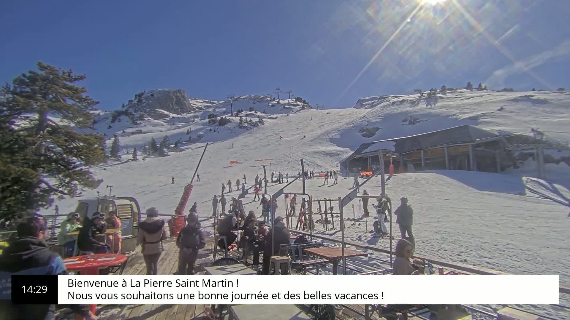 A sunny day at a snow-covered ski resort shows skiers and snowboarders on a slope with a building and lift at the bottom.