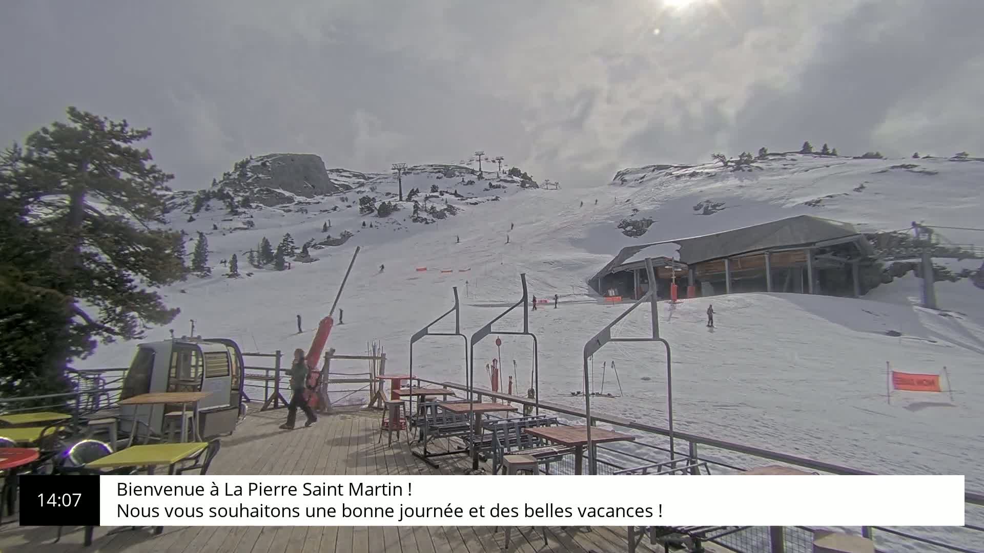A sunny, partly cloudy day on a snow-covered mountain slope with a ski lift and outdoor seating area.