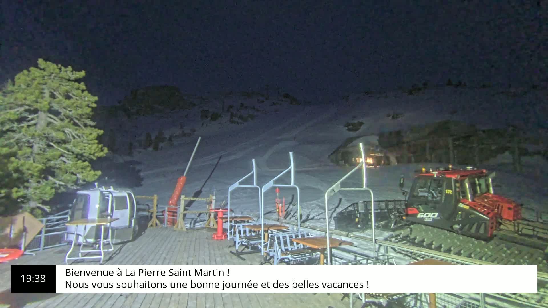 A snowcat is parked near a nighttime ski lift, with several tables and chairs on a wooden deck, and a pine tree to the left.