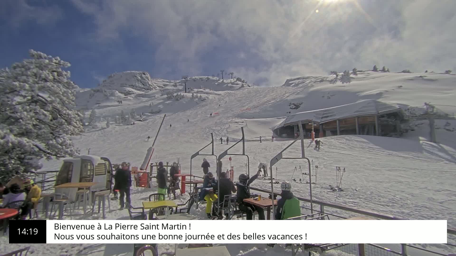 A sunny, snow-covered ski slope with several skiers and a mountainside lodge, featuring a chairlift and people seated at outdoor tables.