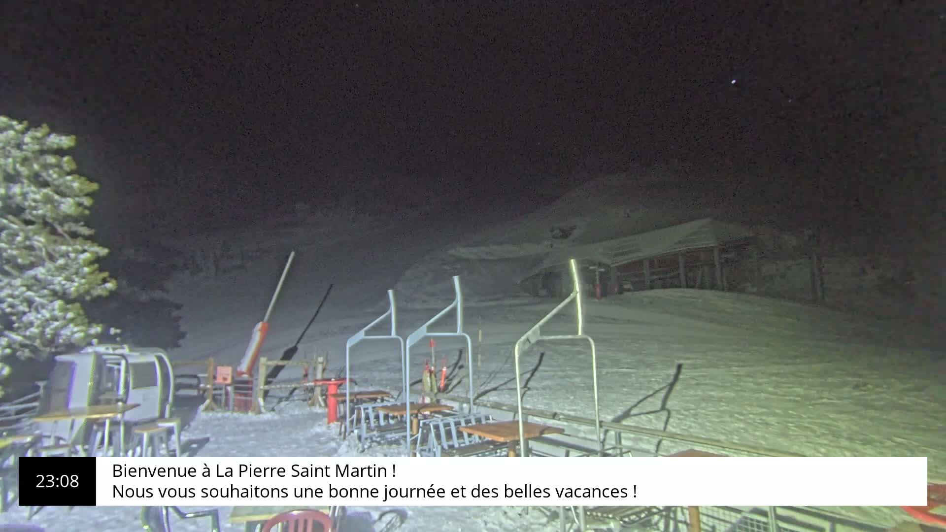 A nighttime scene of a snow-covered ski resort with a chairlift and several tables under a clear, dark sky.