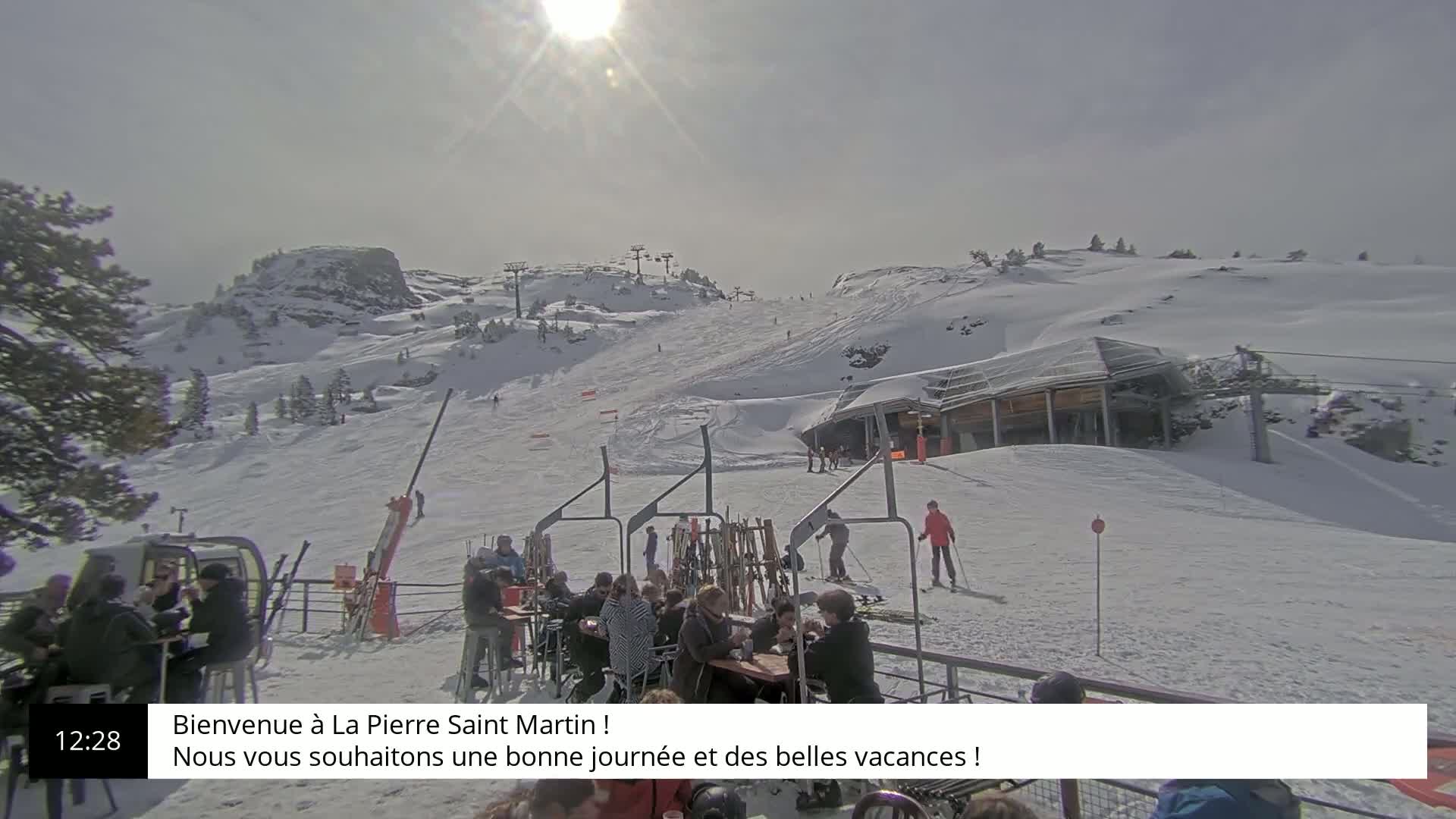 A sunny day at a snow-covered ski resort shows people sitting at outdoor tables near a ski lift and slope with skiers visible.