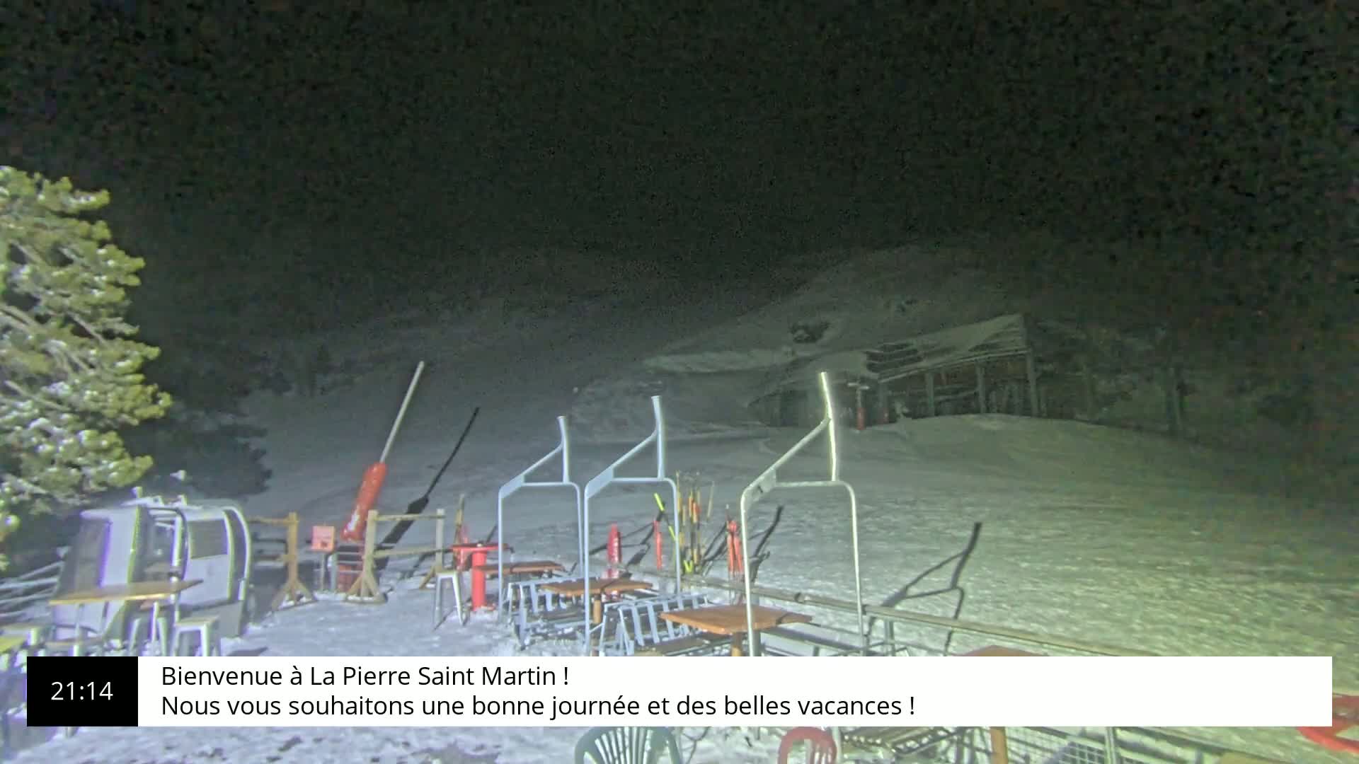 A nighttime, snow-covered ski lift area with several empty tables and chairs is visible under a dark sky.