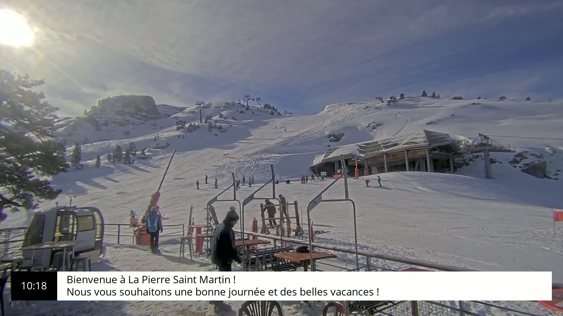 A sunny day at a snow-covered ski resort shows skiers and snowboarders on the slopes, with a lift station and several people near outdoor seating.