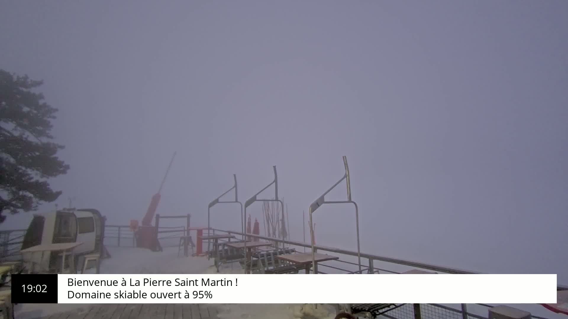 A snowy outdoor scene shrouded in thick fog shows a ski lift station with several empty tables and chairs.