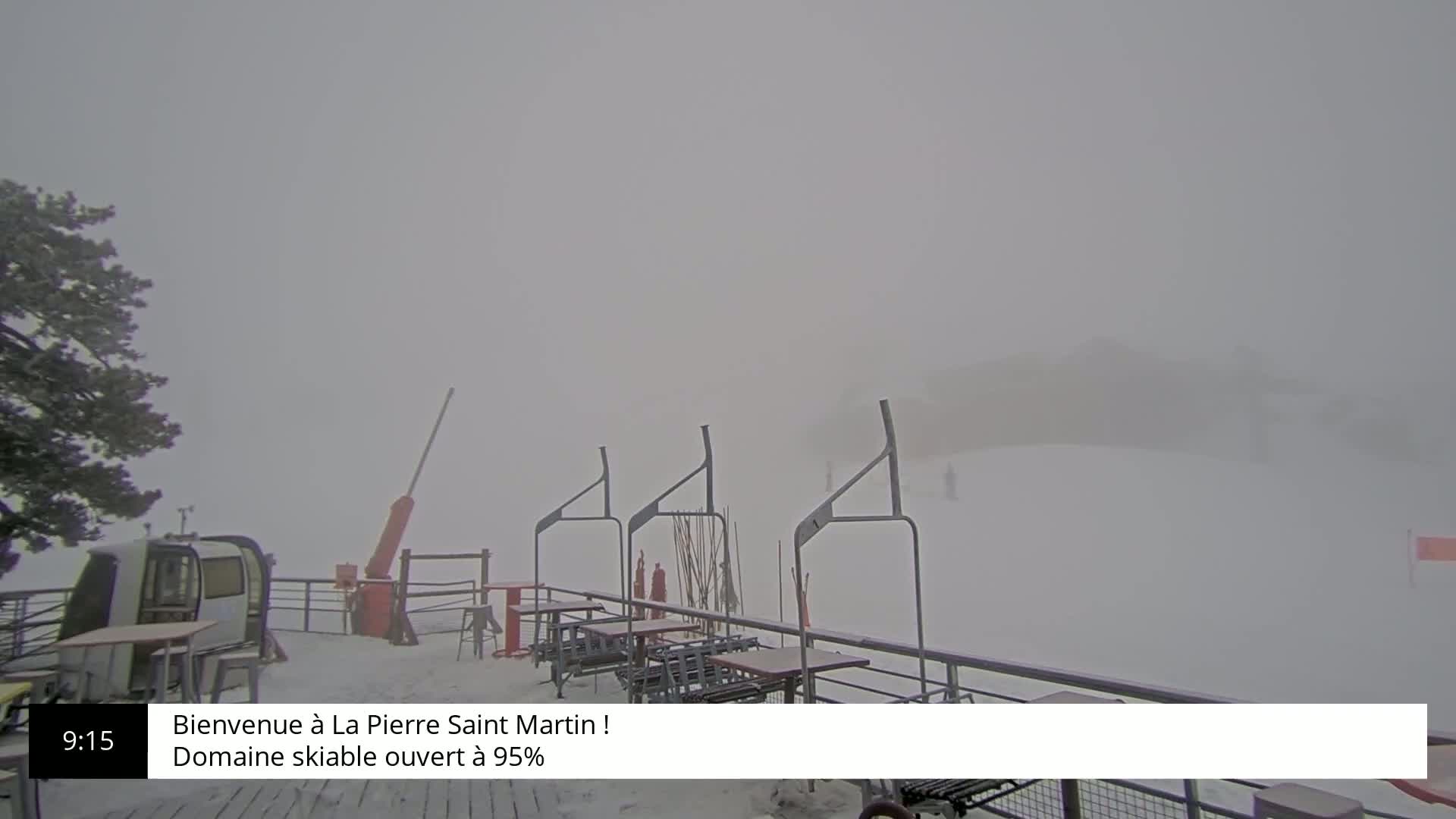 A snow-covered ski lift area is shrouded in heavy fog, with several empty tables and chairs visible near a small building.