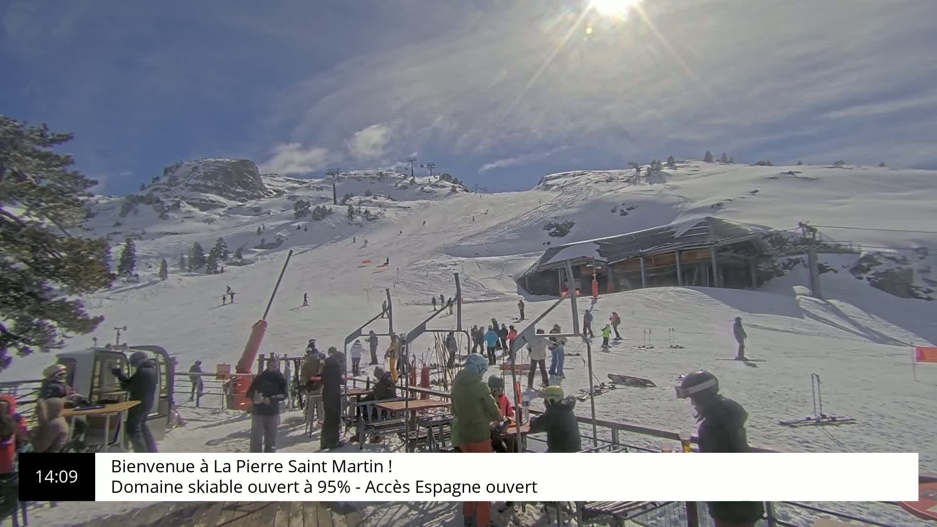 A sunny day at a snow-covered ski resort shows skiers and snowboarders on the slopes with people gathered at outdoor seating near a lift station.