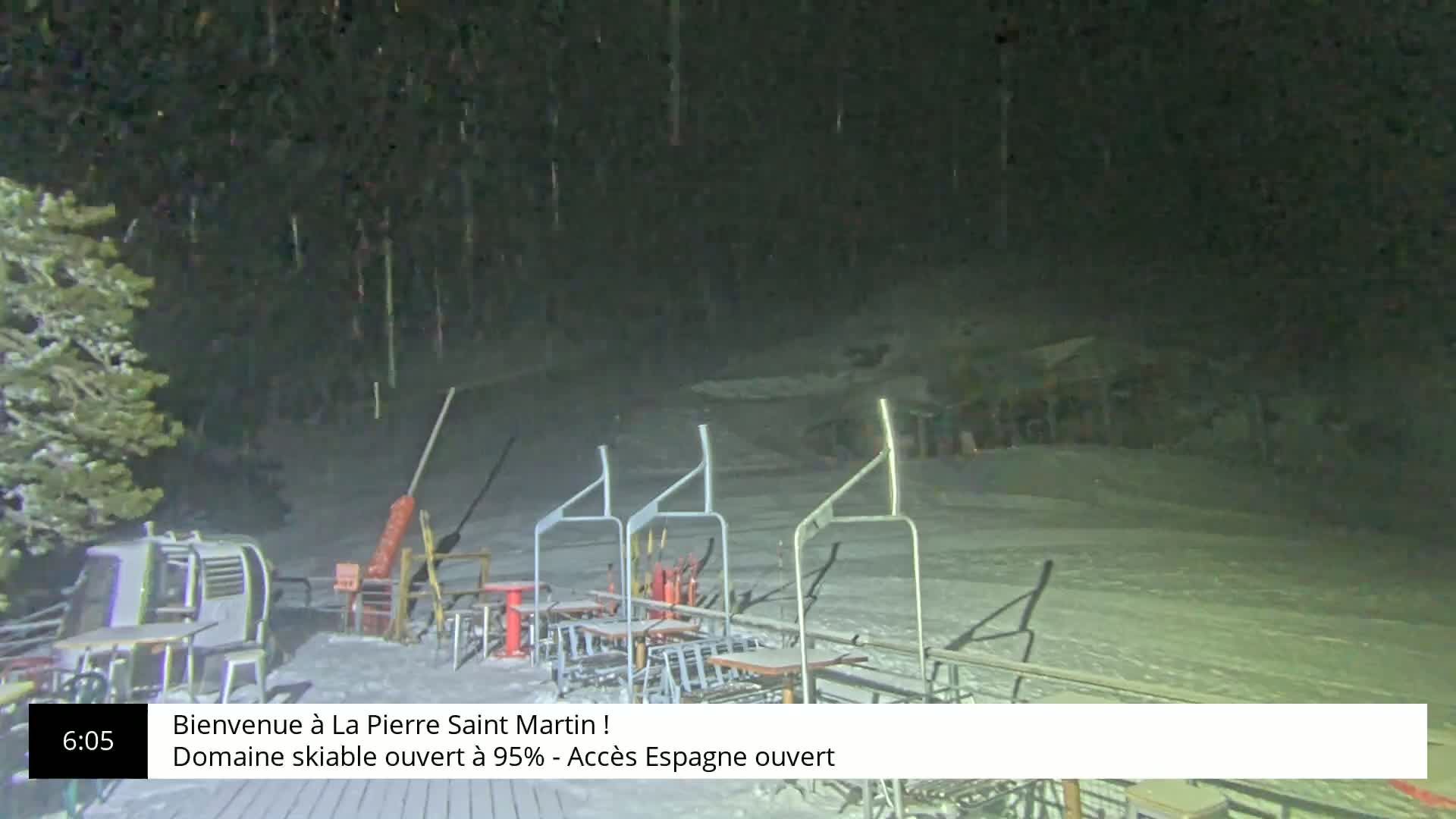 A nighttime view of a snow-covered ski area with several empty chairlifts and tables under a light snowfall.