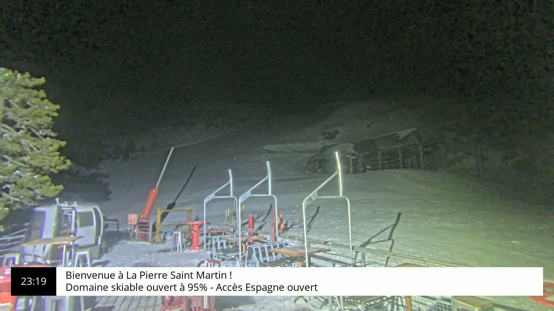A nighttime view of a snow-covered ski resort with a chairlift and several tables under a dark sky.