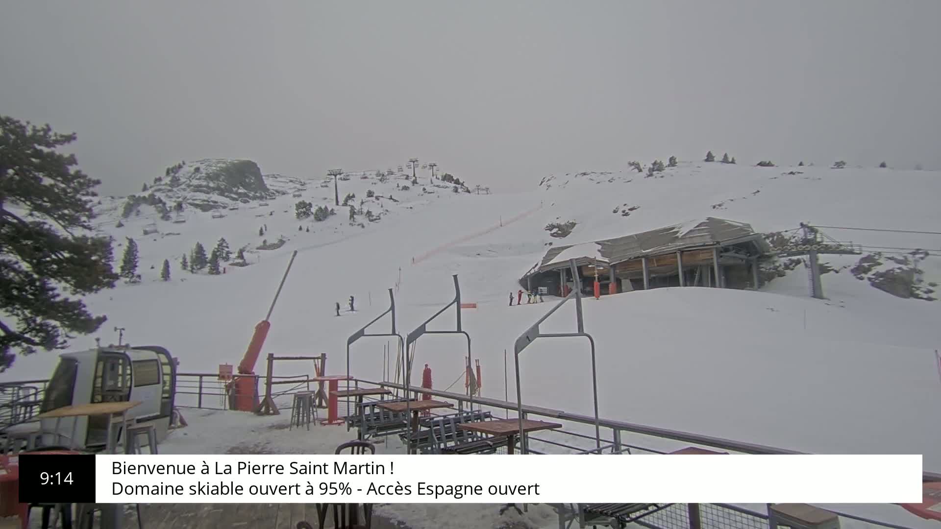 A snow-covered ski slope with a lift station and several skiers visible under an overcast sky.
