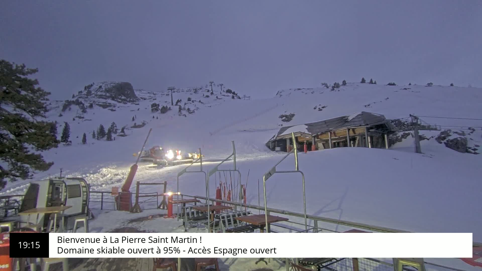 A snow-covered ski resort at dusk, with a snow groomer on a slope and a lift station in the background.