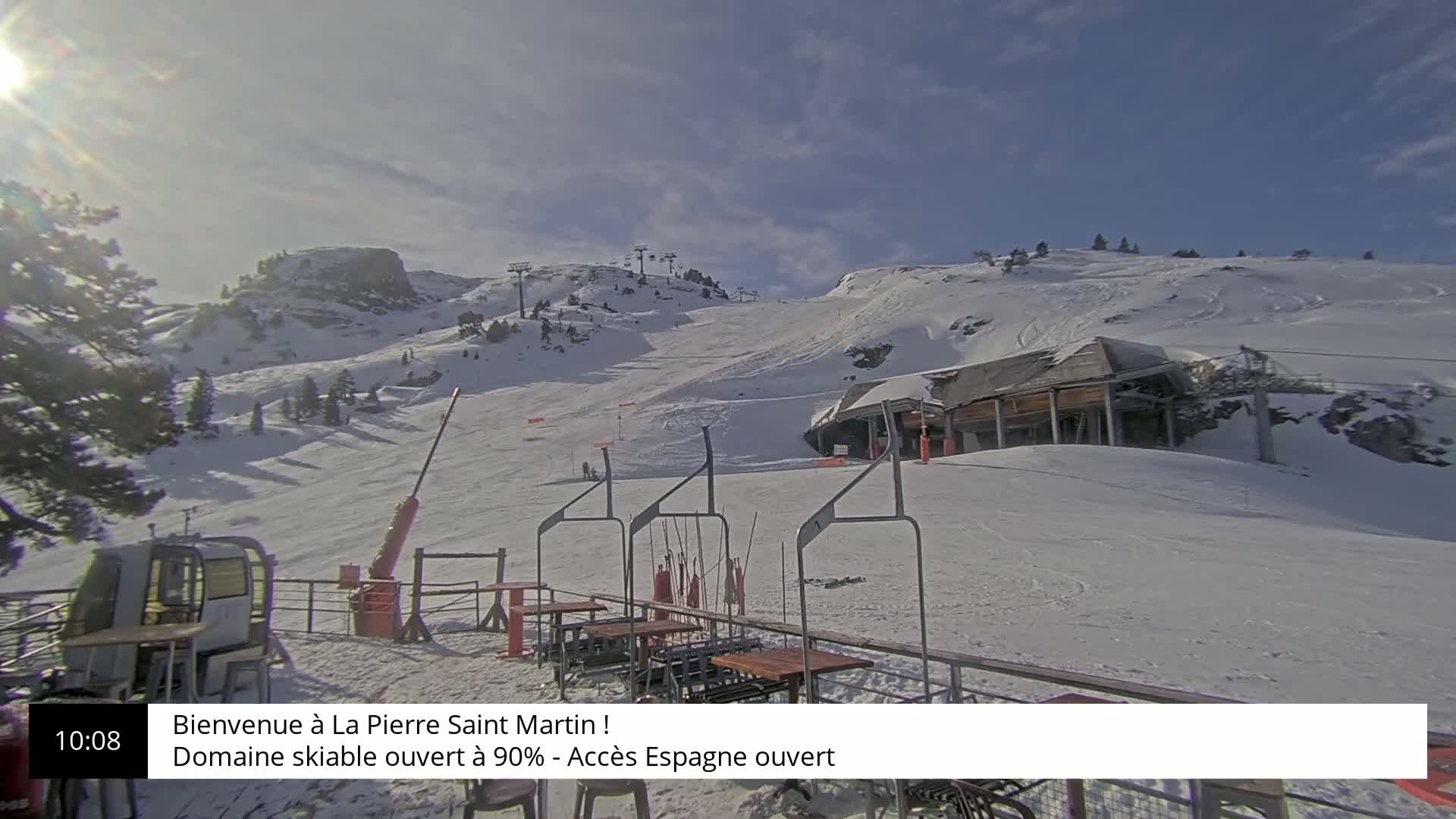 A sunny, snow-covered ski slope with a chairlift and several empty tables sits below a partially cloudy sky.