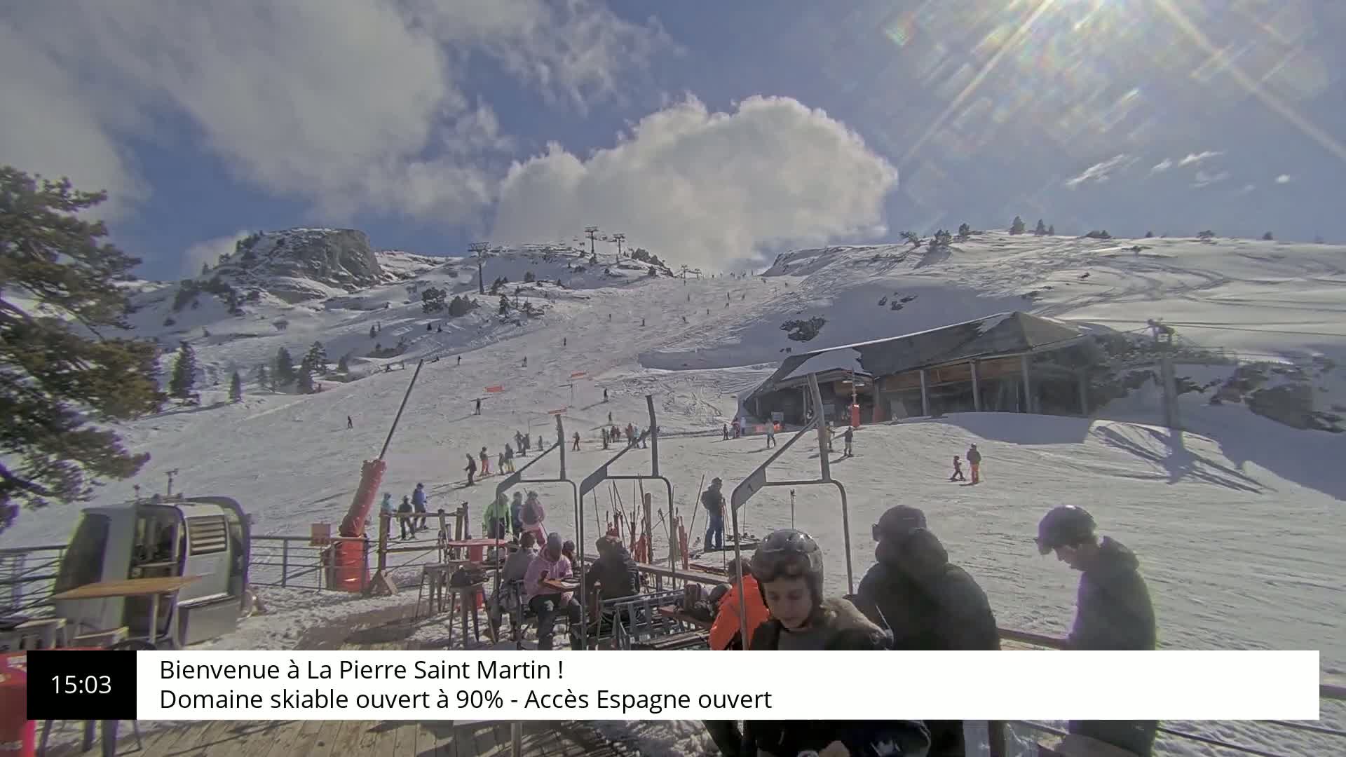 A sunny day at a snow-covered ski resort shows skiers and snowboarders on the slopes, with a chairlift and a building visible in the foreground.
