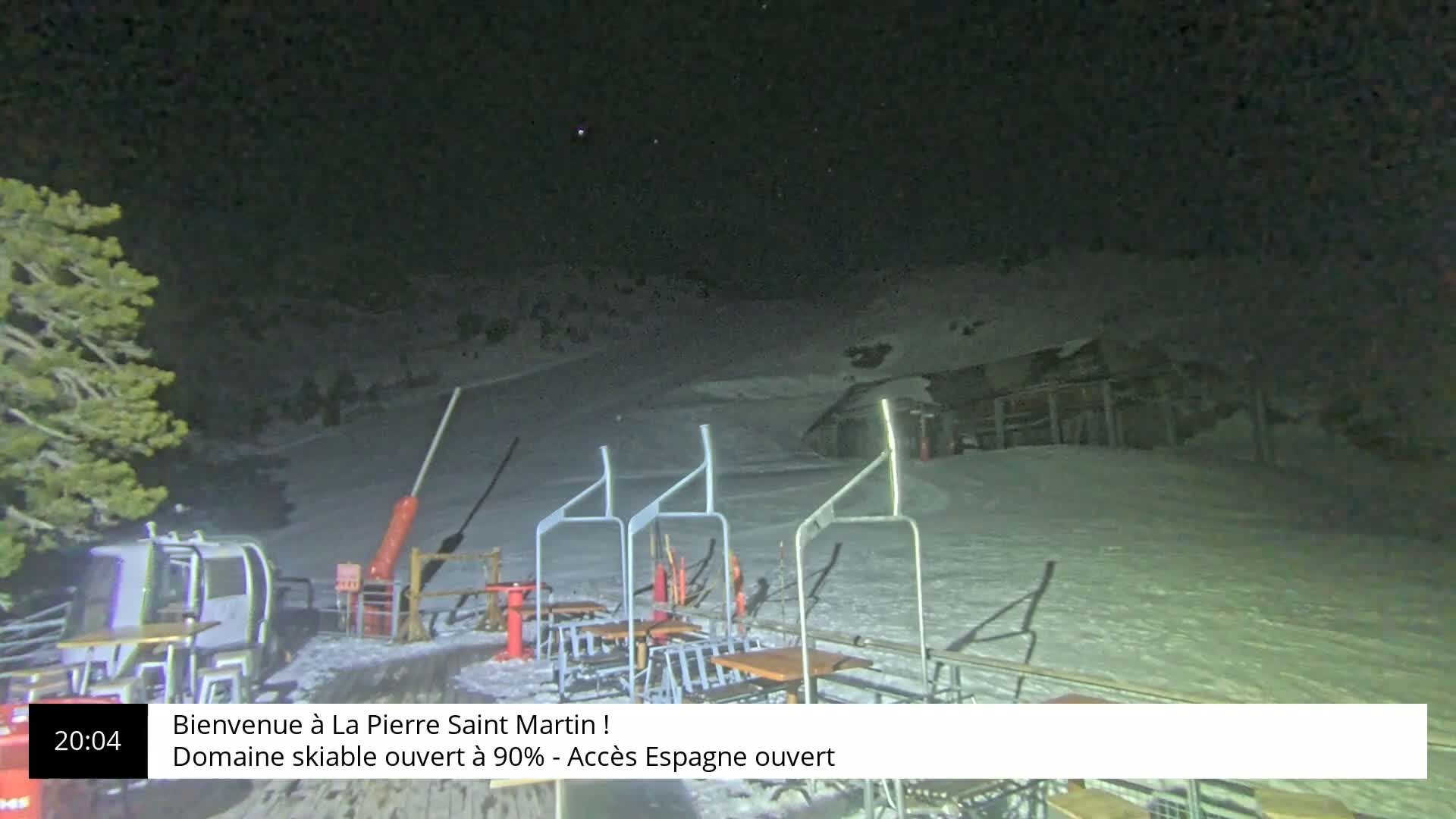 A nighttime view of a snow-covered ski lift area with several empty chairs and tables under a clear, dark sky.