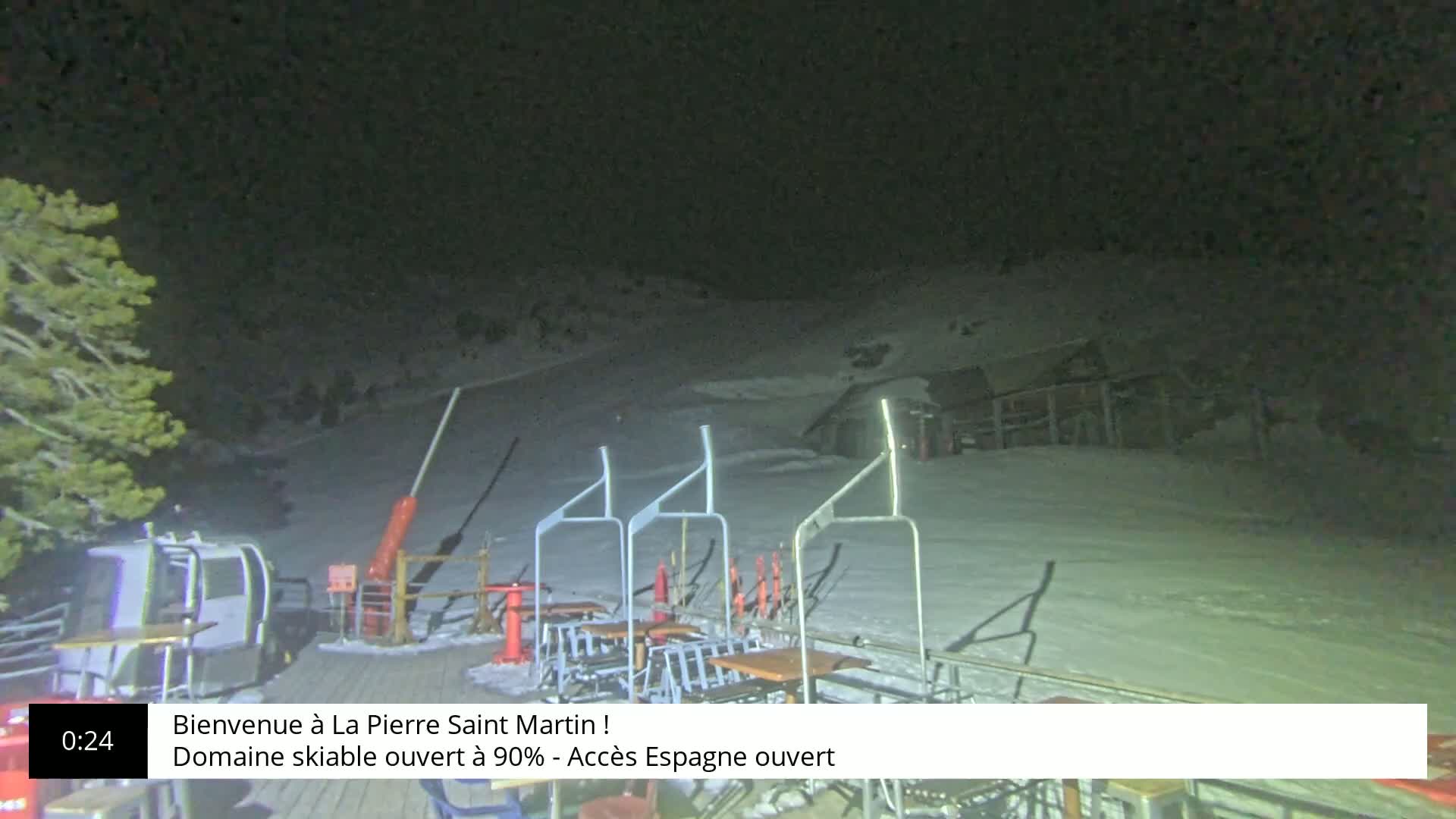 A nighttime scene of a snow-covered ski slope with several empty chairlifts and a few outdoor tables under a dark sky.