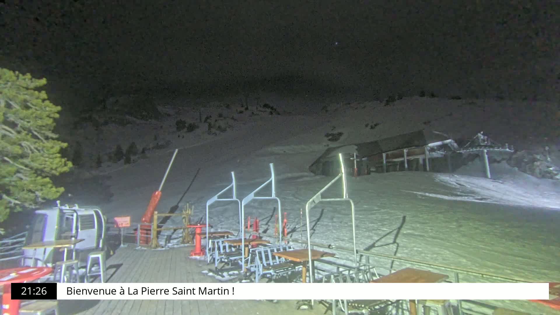 A nighttime view of a snow-covered ski slope with a chairlift and outdoor seating area illuminated under a dark sky.