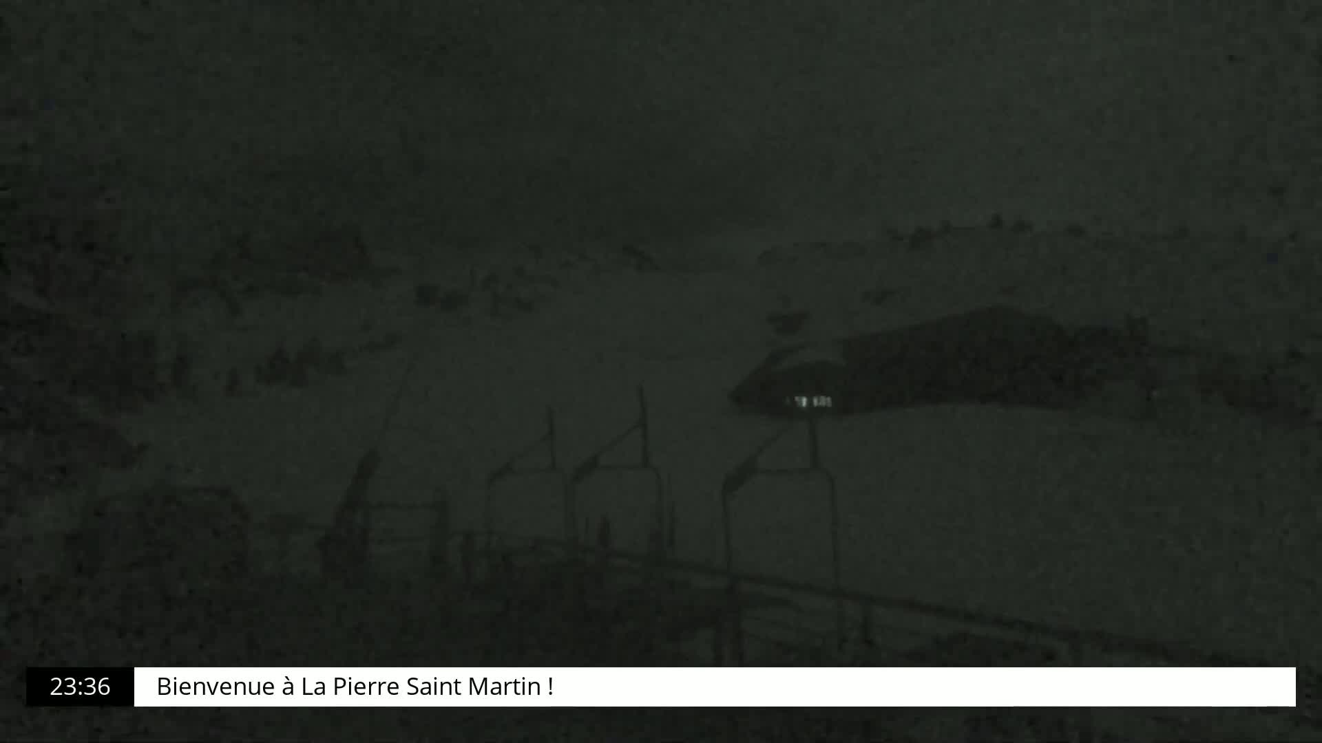 A nighttime, snow-covered scene shows a dark landscape with indistinct shapes suggesting ski lifts and possibly a building under heavy snowfall.