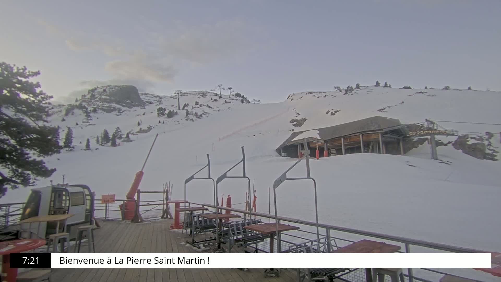 A snow-covered ski slope with a ski lift and a building, viewed from a wooden deck with outdoor seating under an overcast sky.