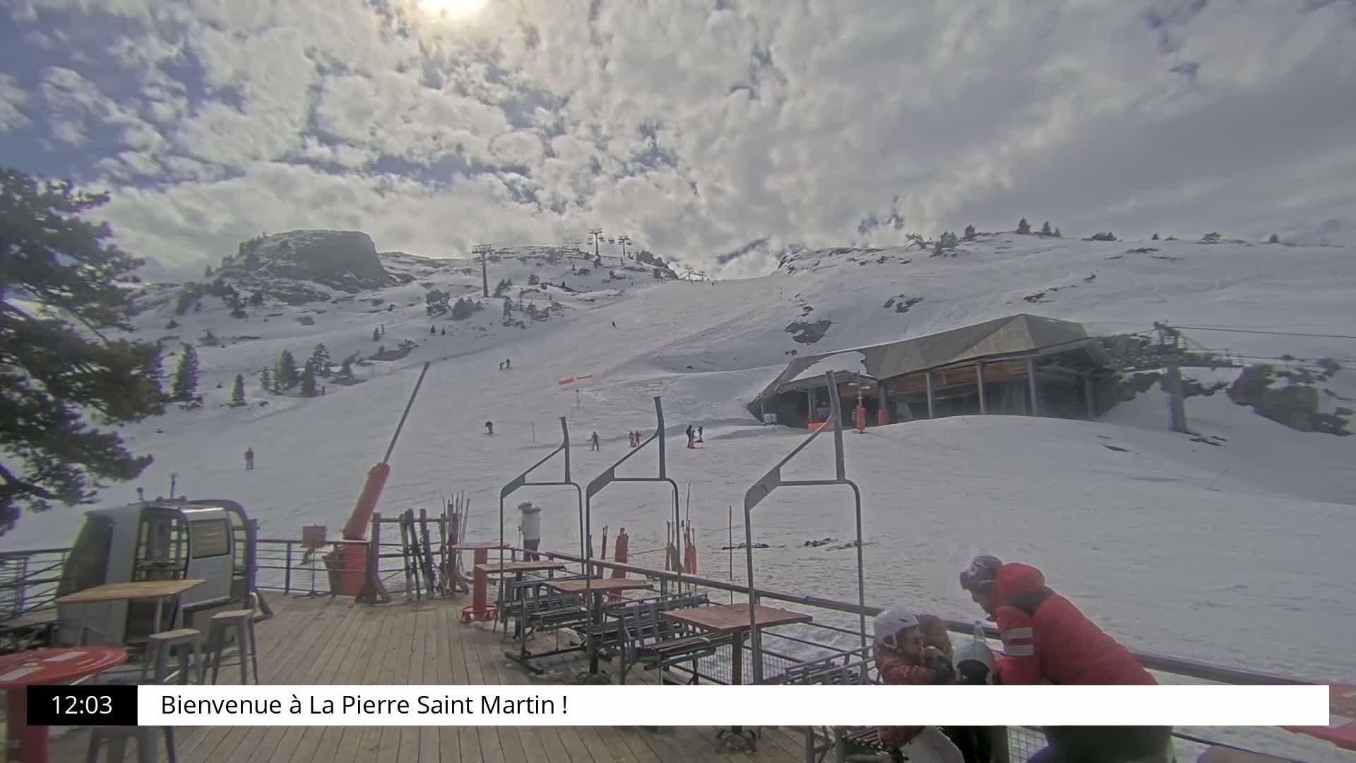 A partially sunny, cloudy day at a snow-covered ski resort shows a ski lift, skiers, a building, and outdoor seating on a deck.