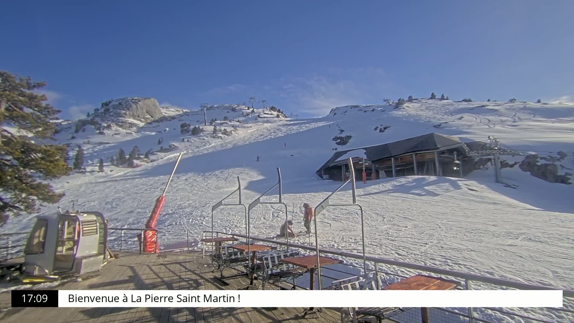 A sunny winter scene shows a snow-covered ski slope with a chairlift, a small building, and several people near a deck with outdoor seating.