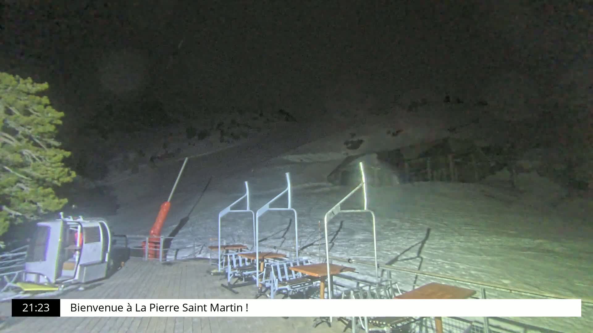 A nighttime view of a snow-covered ski slope with several empty chairlift stations and tables, illuminated by artificial light.
