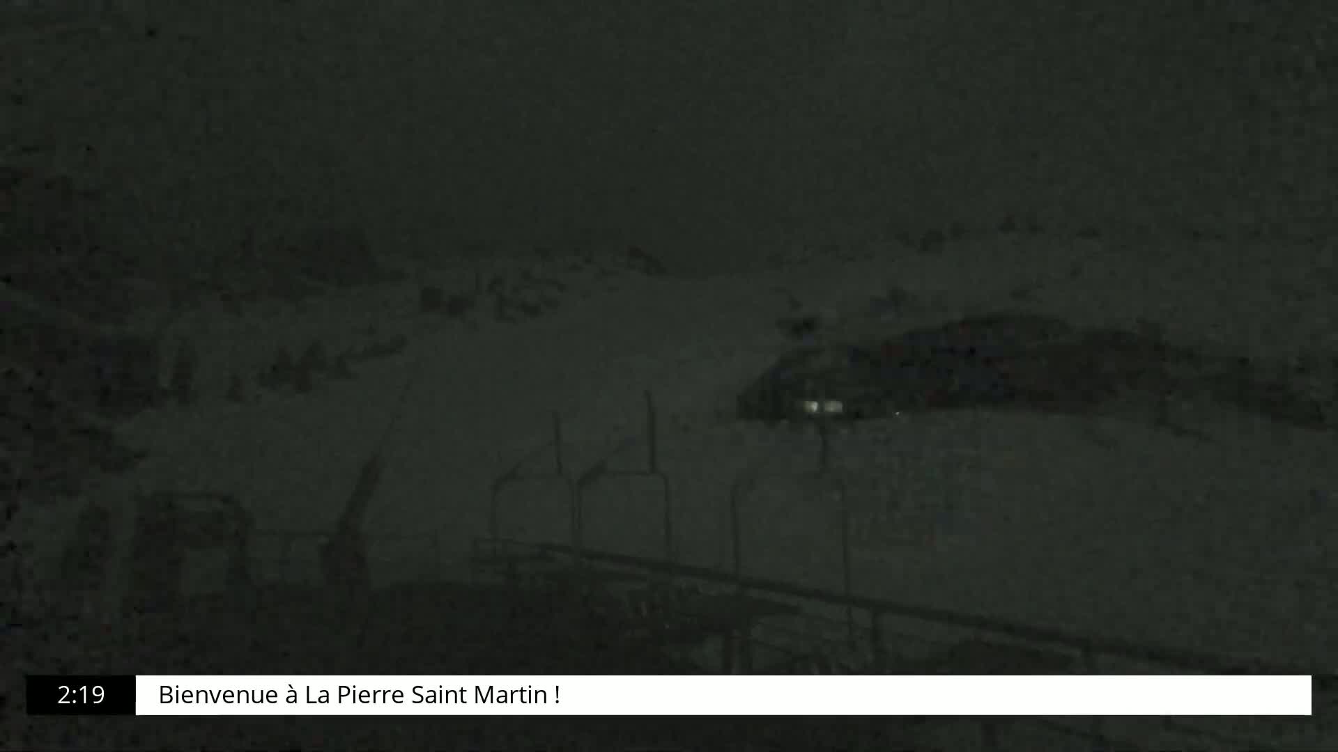 A dark, snowy scene at night shows a few indistinct shapes in the distance, possibly vehicles, with some metal structures in the foreground.