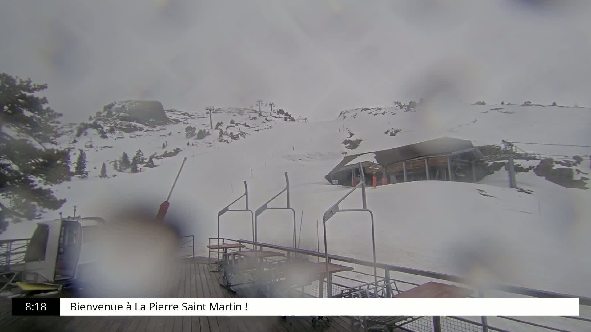 A snowy ski slope is viewed from a deck on an overcast day, with a ski lift and building visible in the distance.