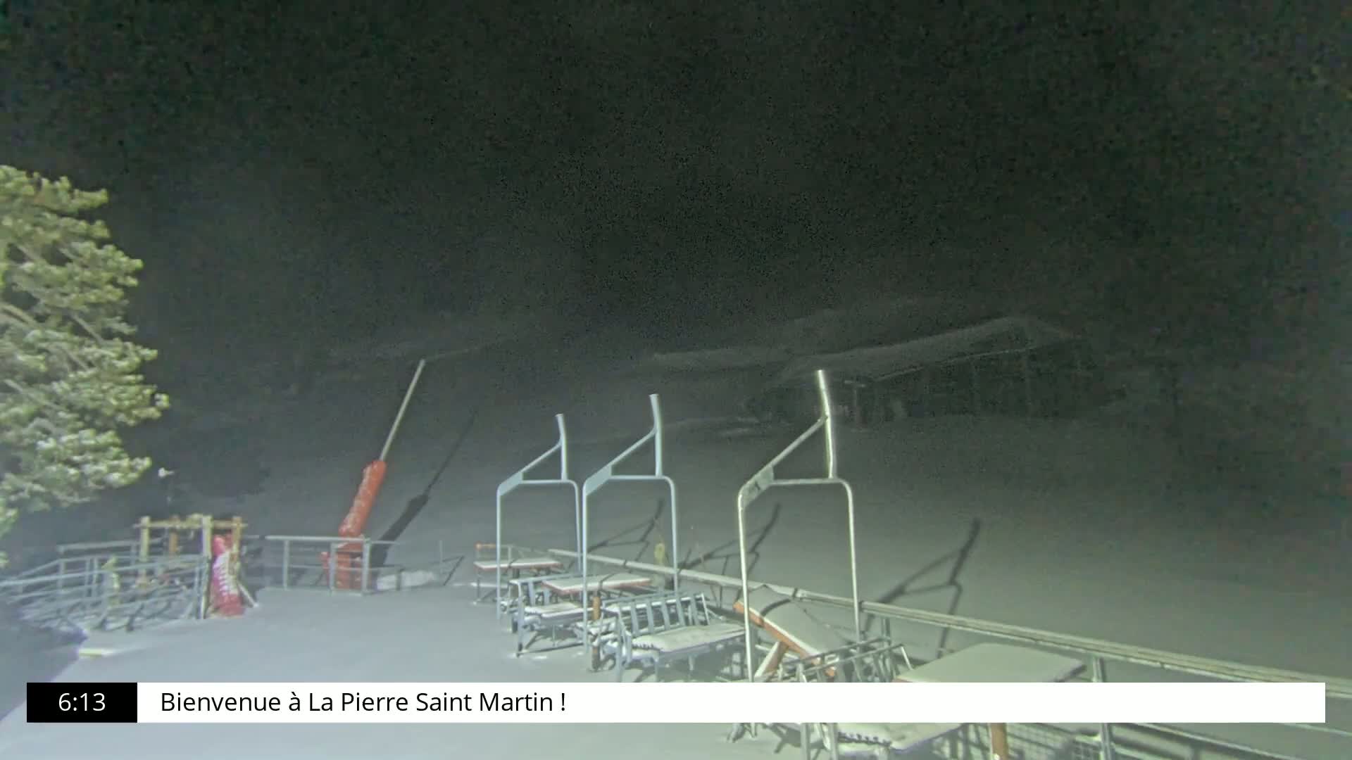 At night, under a dark sky, several empty ski lift chairs are shown covered in snow, with a snow-covered slope visible in the background.