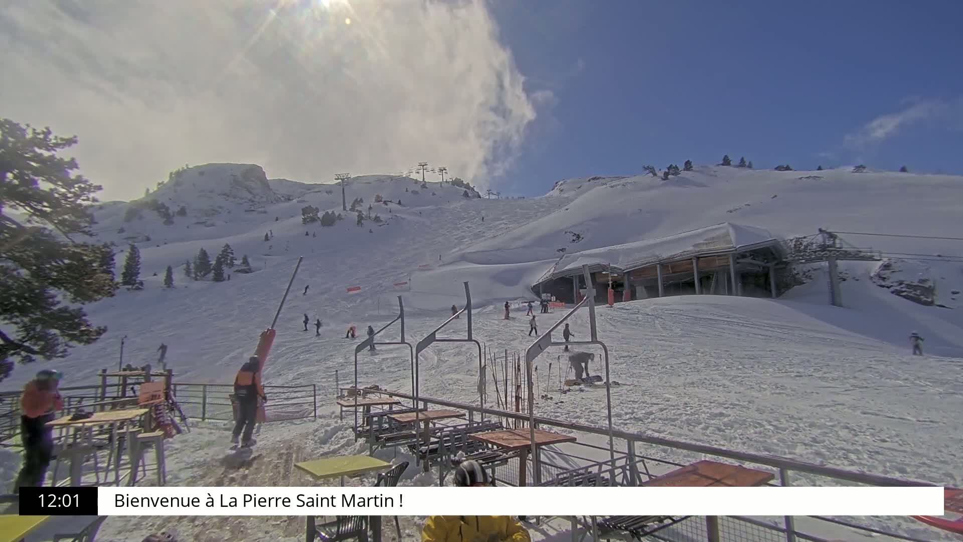 A sunny, snow-covered ski slope with a ski lift and several skiers visible, along with outdoor seating at the bottom of the hill.
