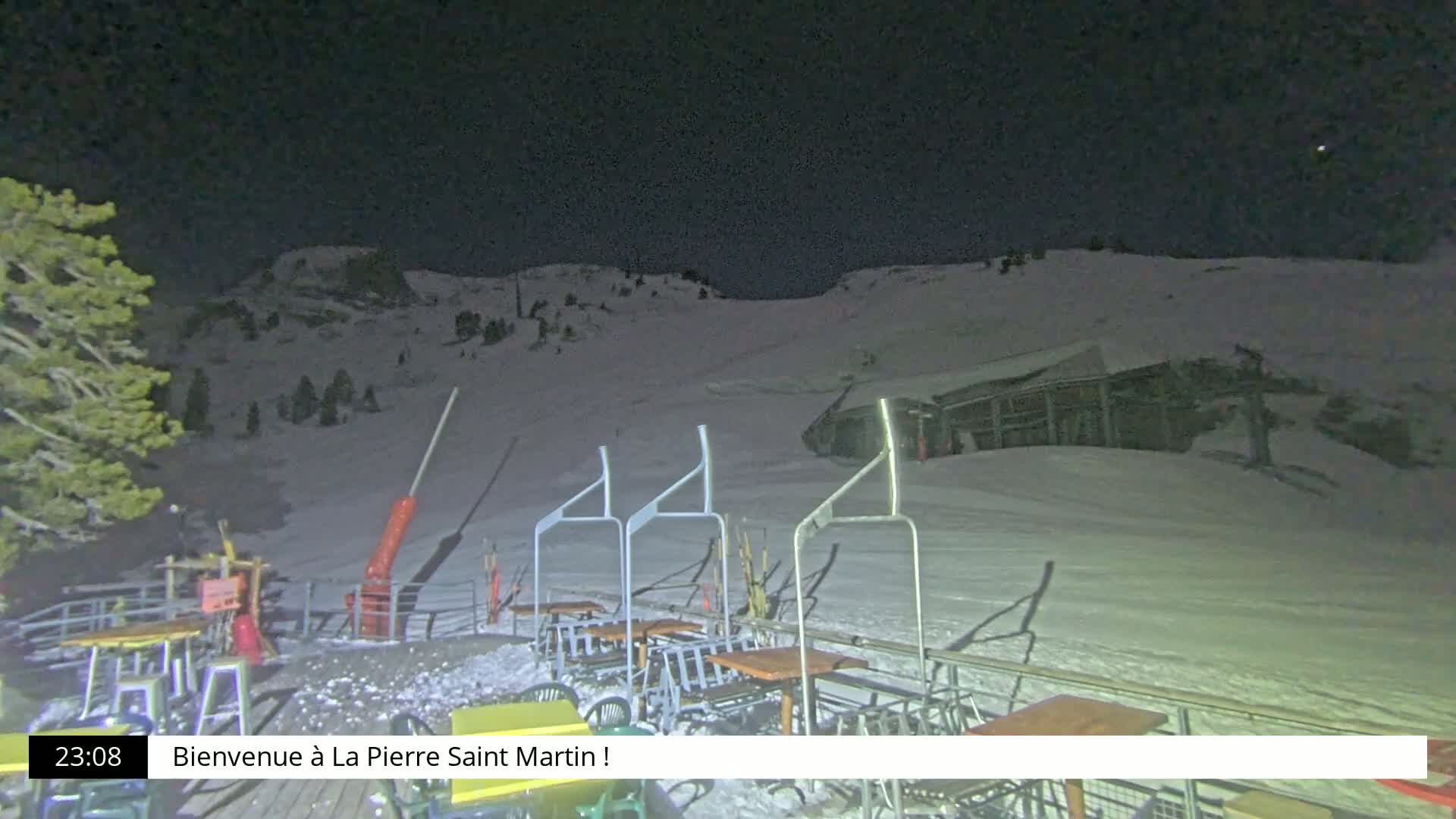 A nighttime view of a snow-covered ski slope with a chairlift and outdoor seating area under a dark, clear sky.