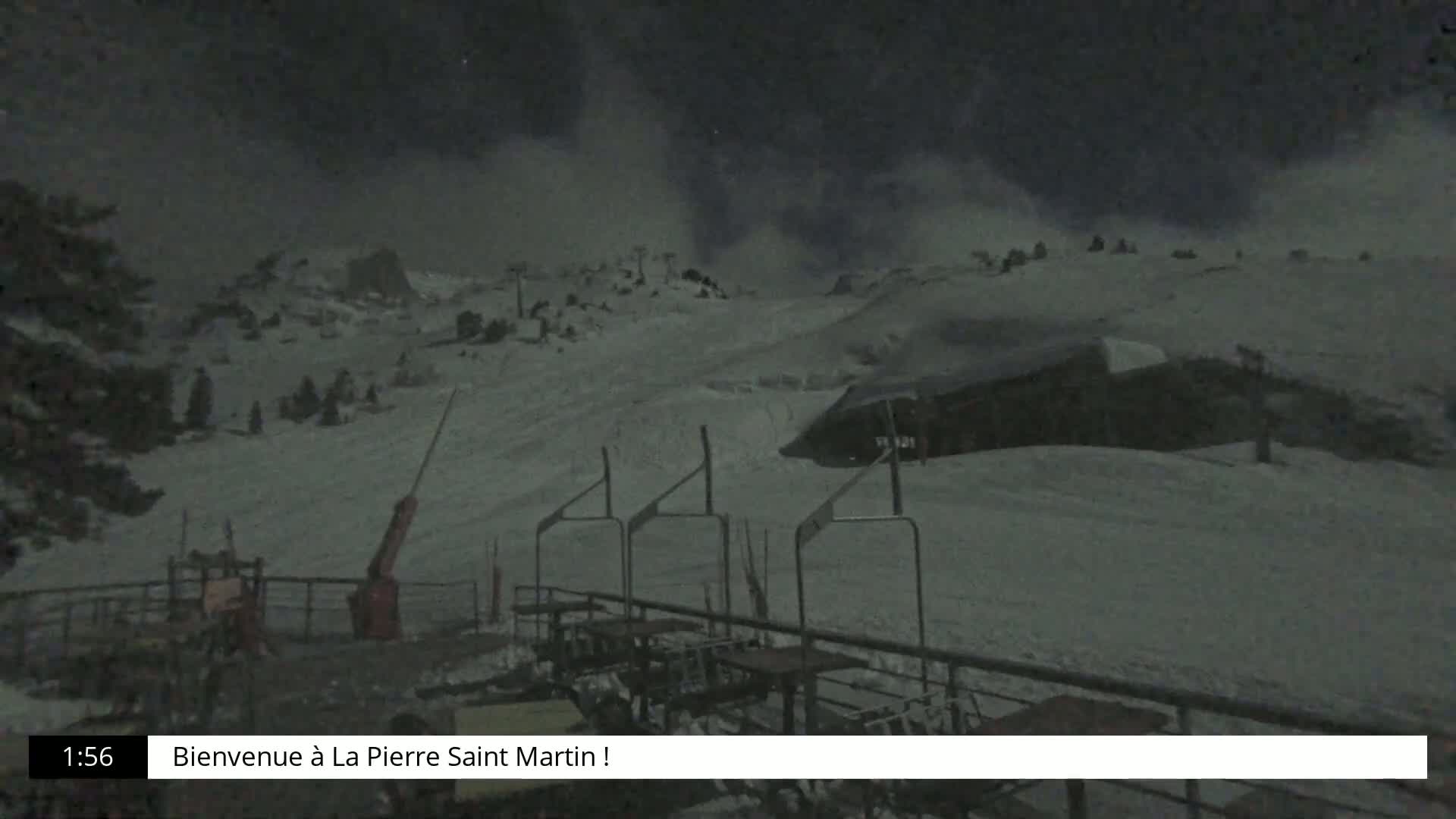 A snow-covered ski slope at night, under a dark, cloudy sky, is viewed from a ski lift base with several empty chairs and tables.