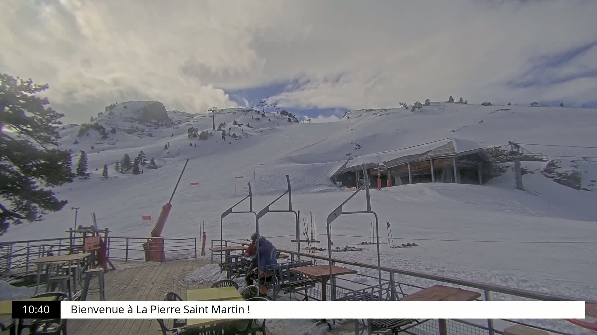A mostly cloudy day at a snow-covered ski resort shows a ski lift and a building at the bottom of a hill with a few people sitting at outdoor tables.