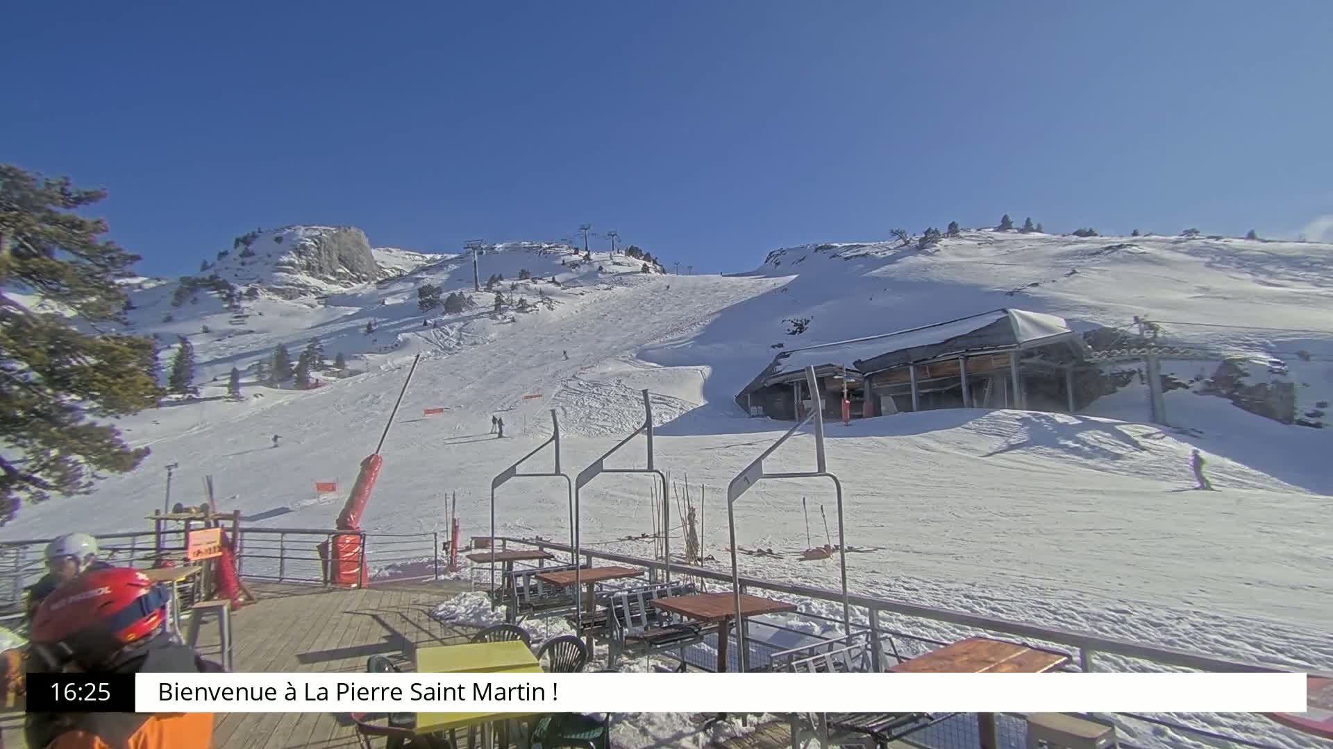 A sunny, snow-covered mountain slope with a ski lift and outdoor seating area is visible.