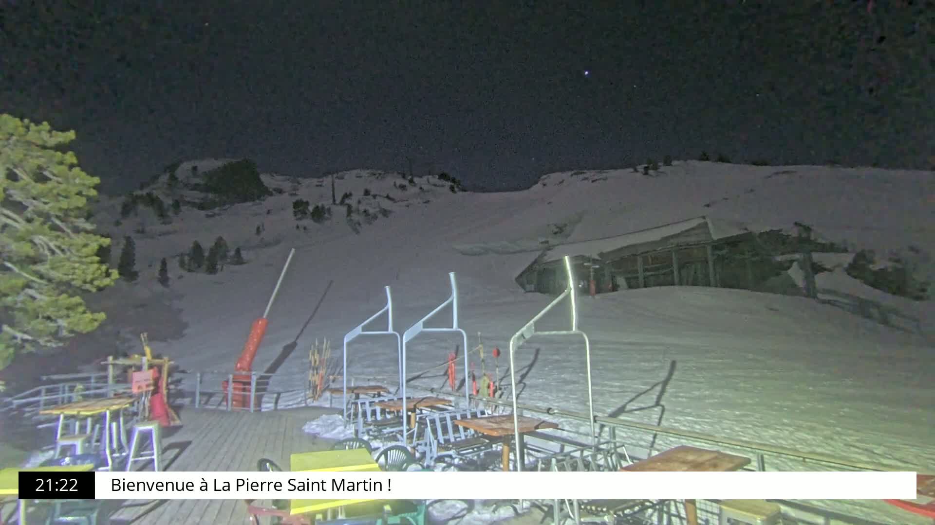 A nighttime view of a snow-covered ski slope with a small building and empty chairlift supports on a deck with tables and chairs.