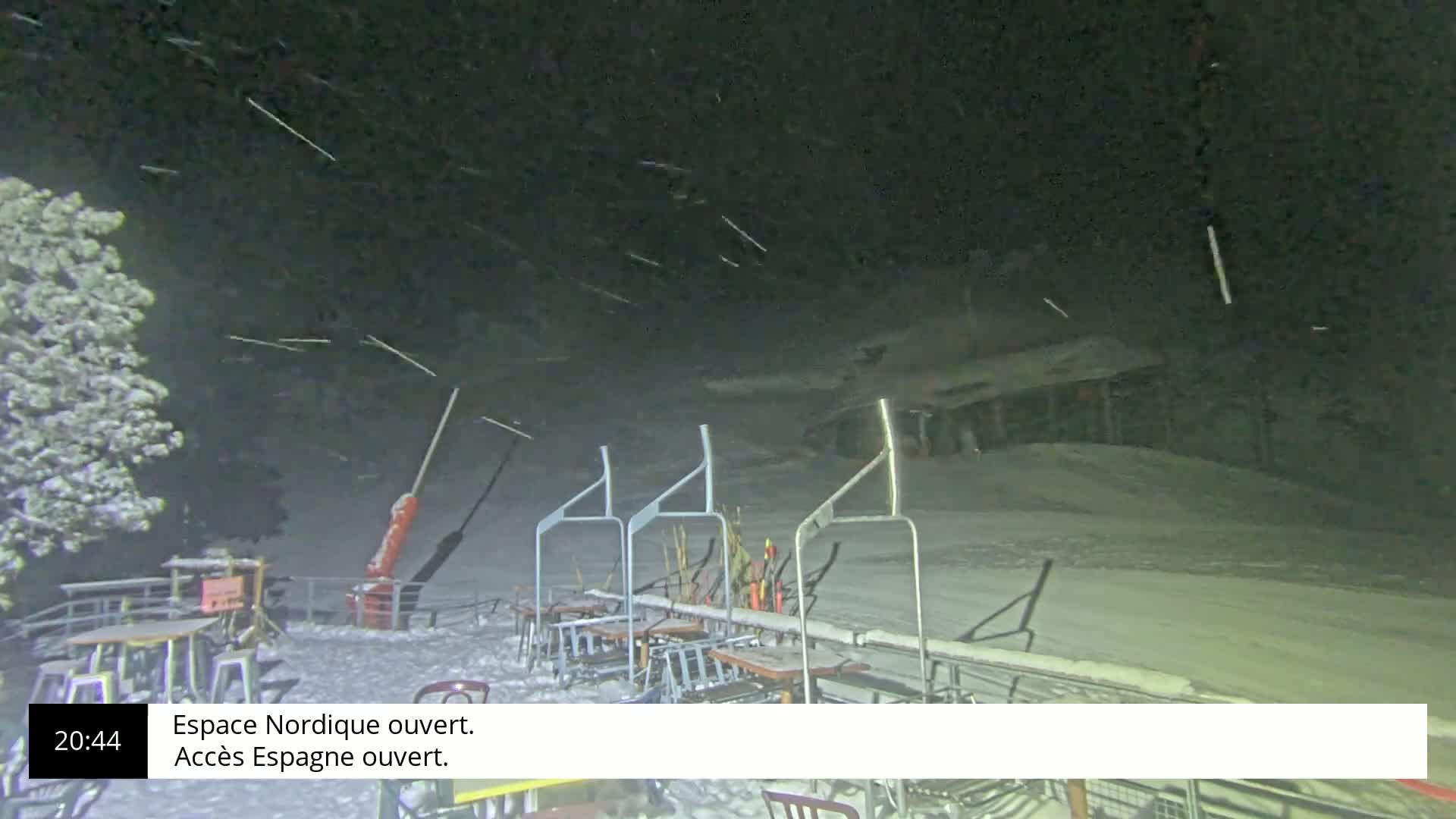 A nighttime snow flurry falls over a snow-covered ski lift and outdoor seating area.