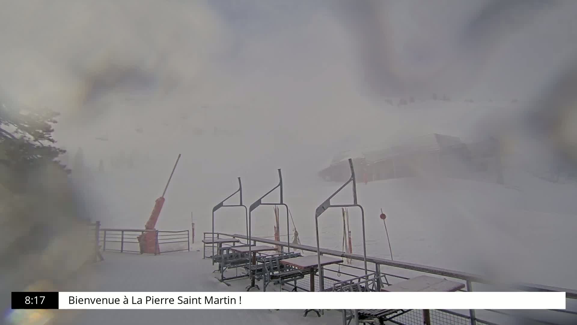 A snowy outdoor scene shows several empty chairlift stations and tables on a patio, with heavy snow obscuring the view of the surrounding area.
