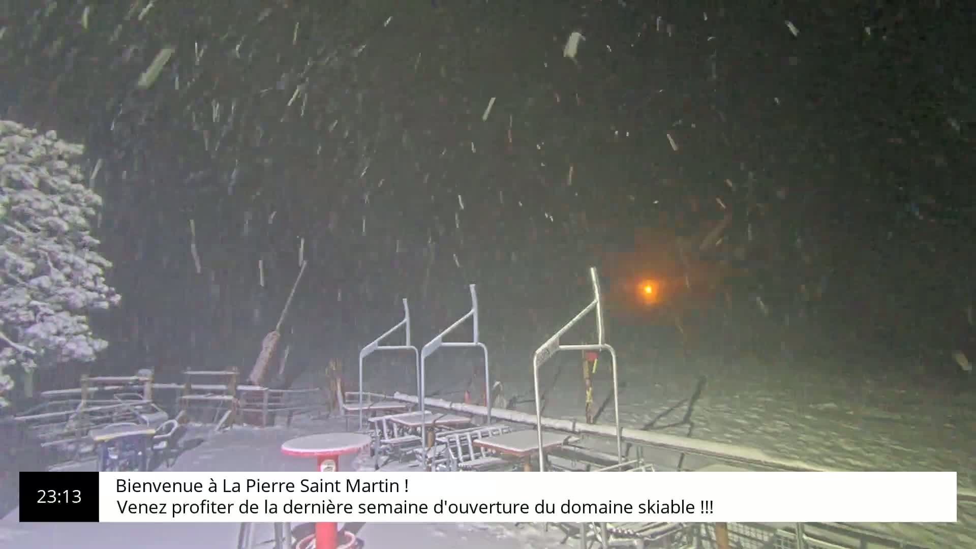 A nighttime scene of snow falling heavily over an outdoor ski lift area with several empty tables and chairs covered in snow.