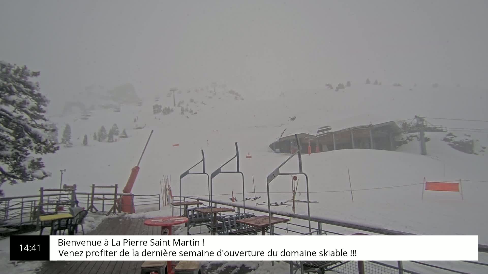 A snowy mountain ski resort is viewed from a deck, with a heavy snowfall obscuring the distant mountains and ski lifts.