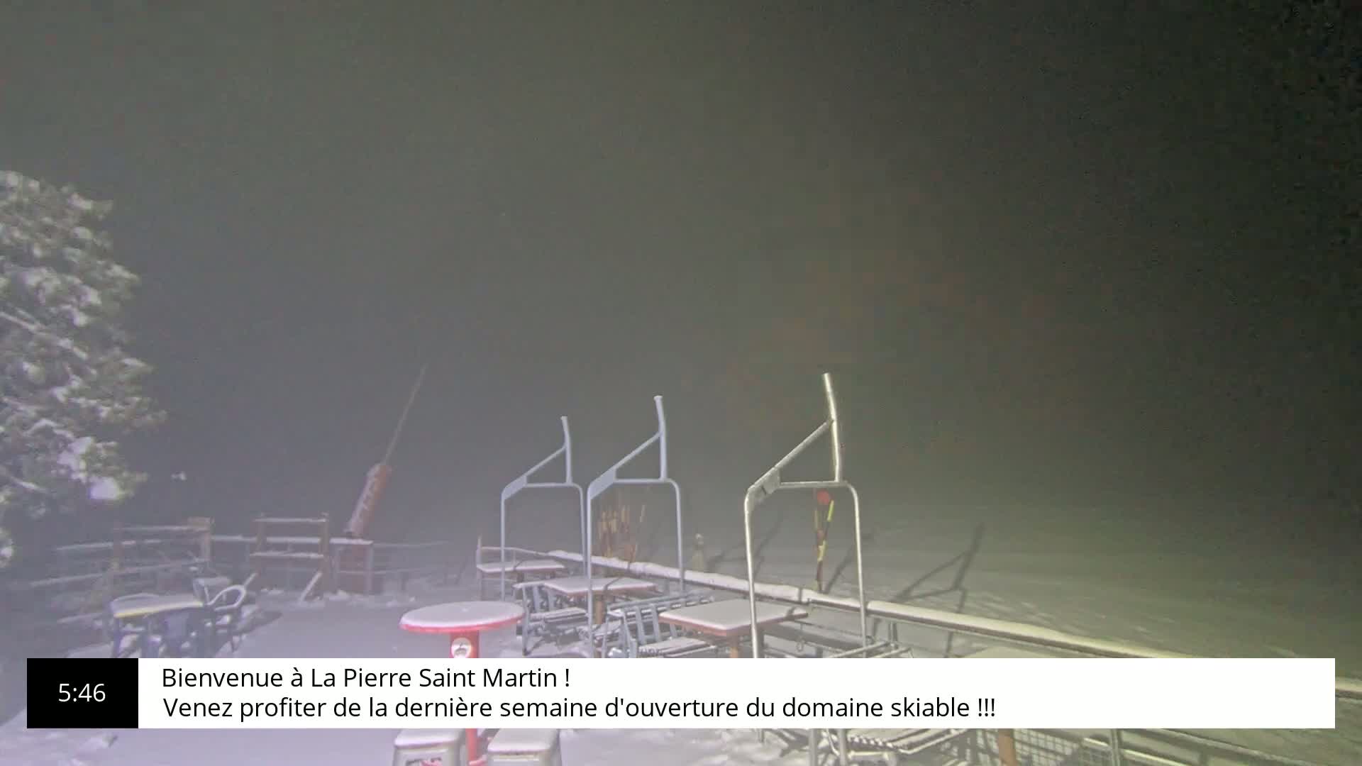 A snow-covered outdoor area at night, shrouded in fog, features several empty tables and chairs near a ski lift.