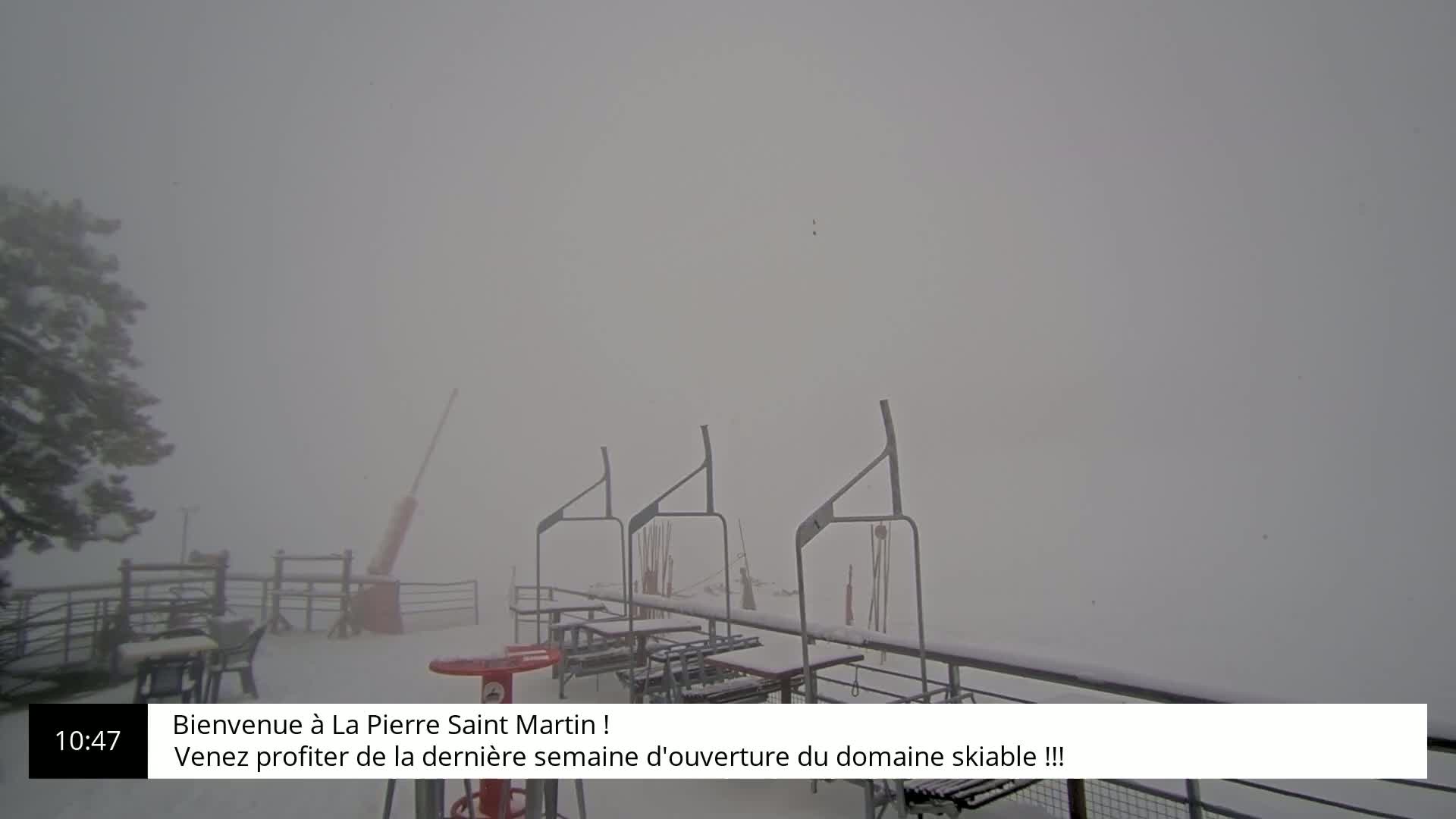 A snow-covered outdoor scene shows several empty picnic tables and chairlifts under heavy, blowing snow.