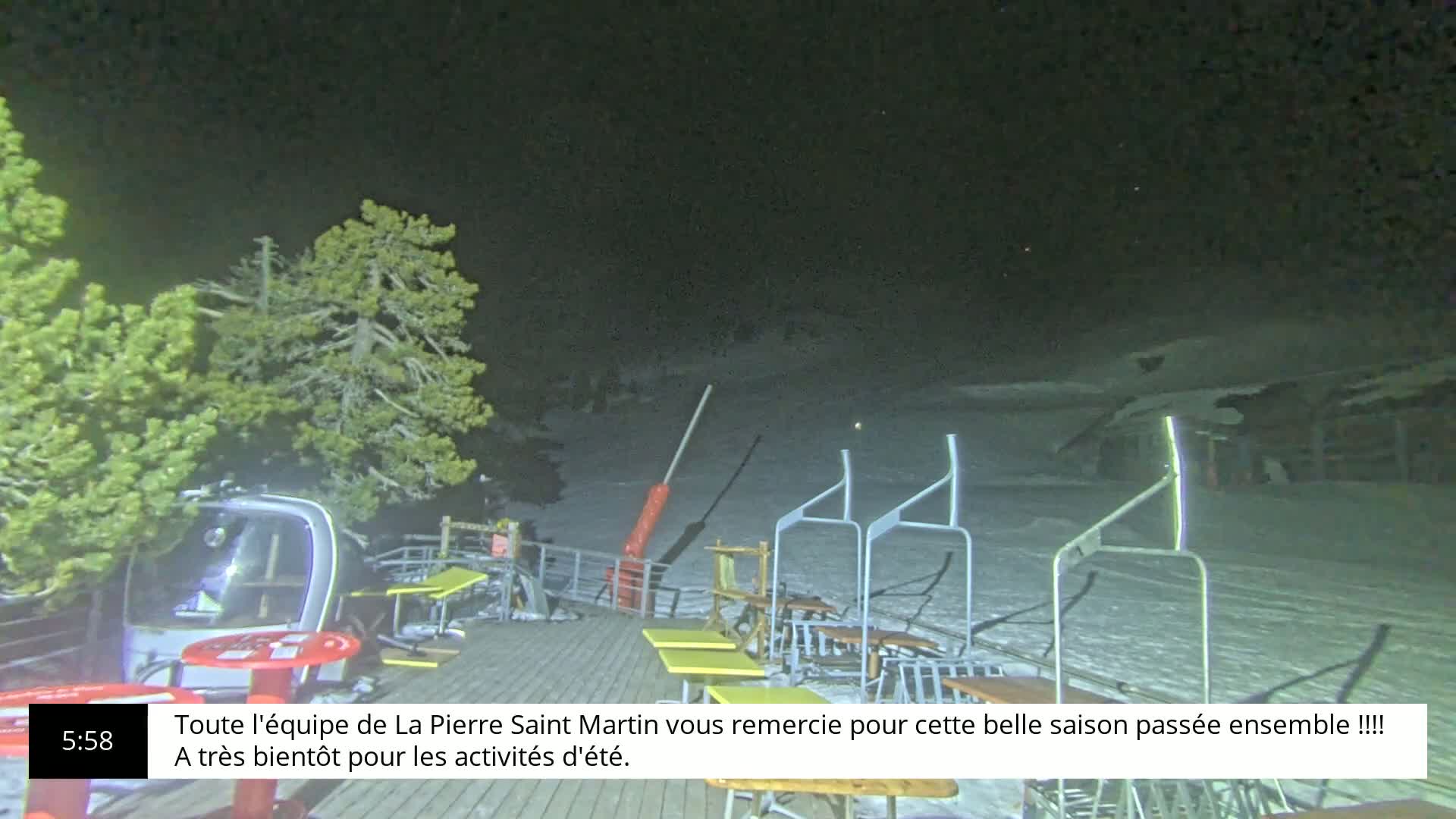 A nighttime scene shows a snow-covered area with several empty chairlift supports, a few tables and chairs on a wooden deck, and some evergreen trees under a clear, dark sky.