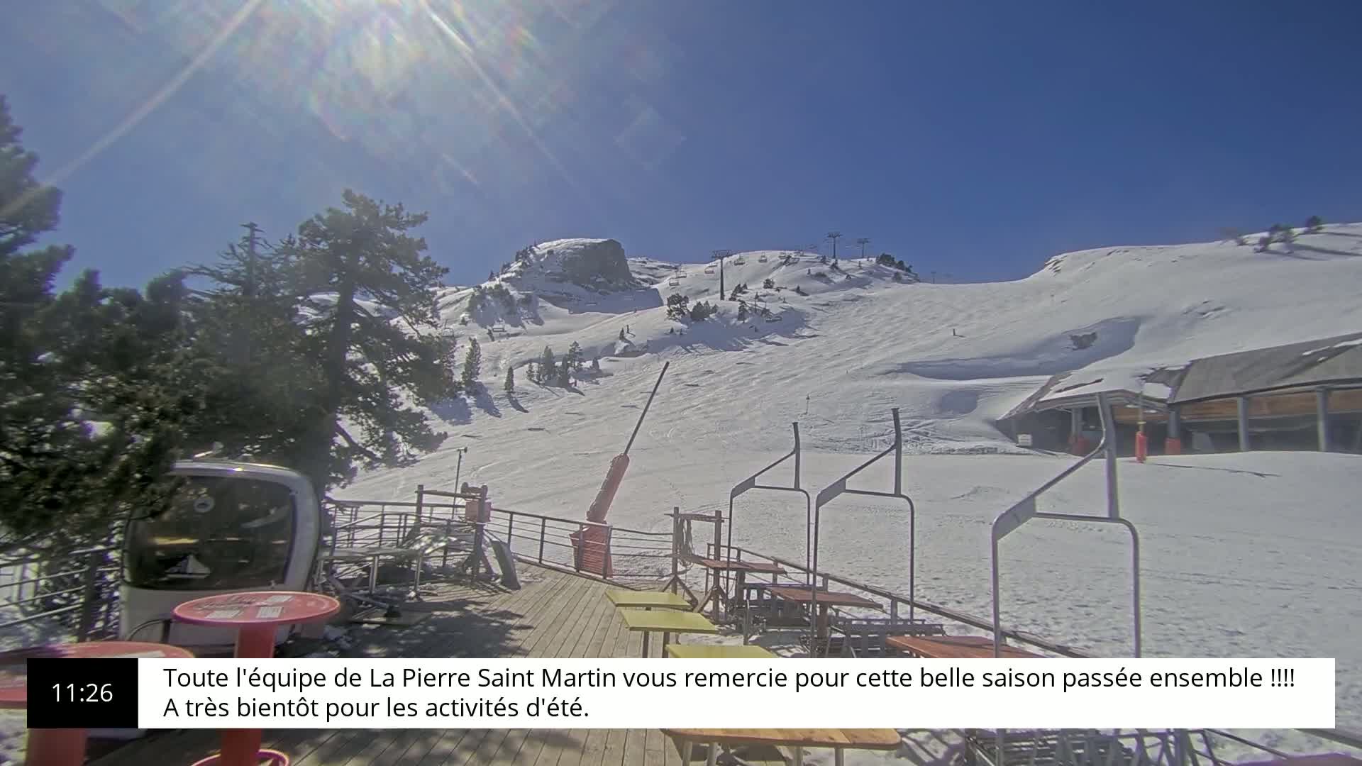 A sunny day at a snow-covered ski resort shows a mostly empty ski slope with a chairlift and several outdoor tables.
