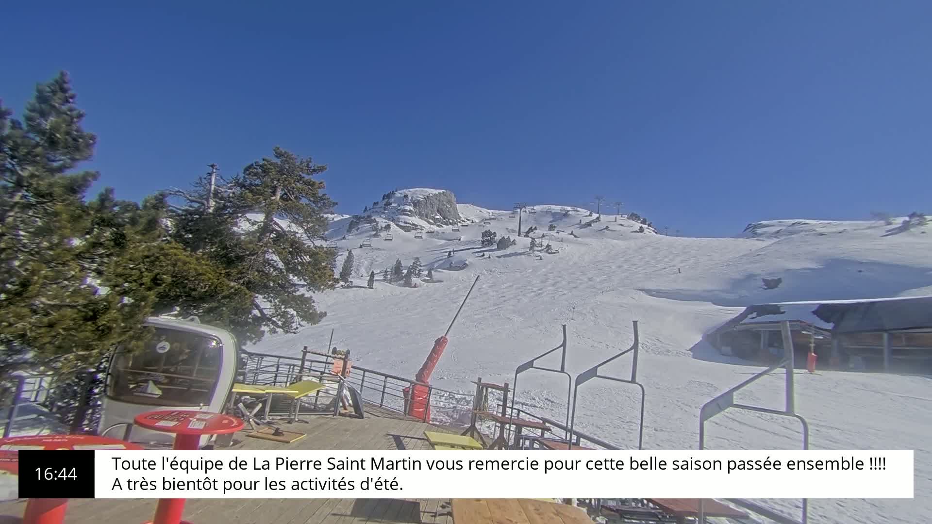 A snow-covered ski slope is visible from a mountaintop patio on a clear, sunny day.