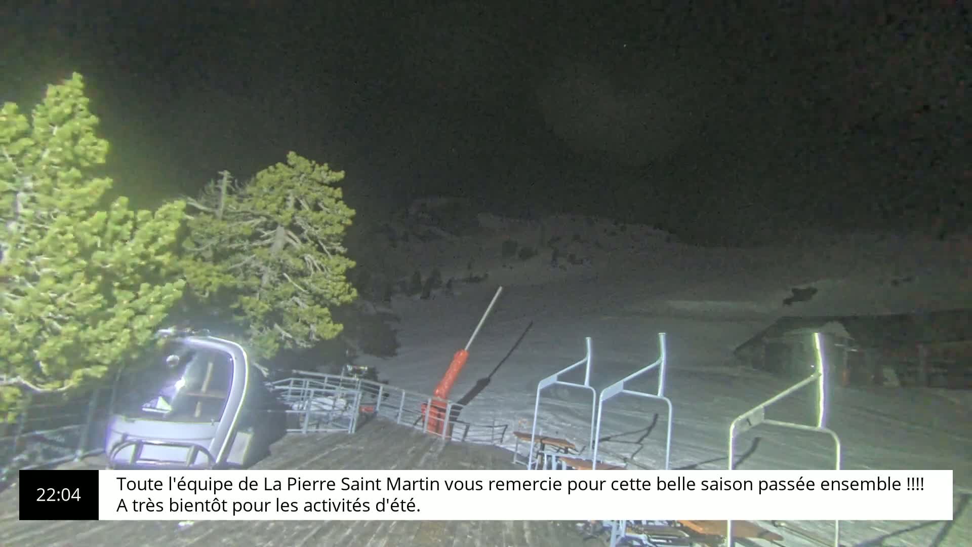 A nighttime scene shows a snow-covered ski lift with a chairlift car at the bottom, illuminated by artificial light under a dark sky.